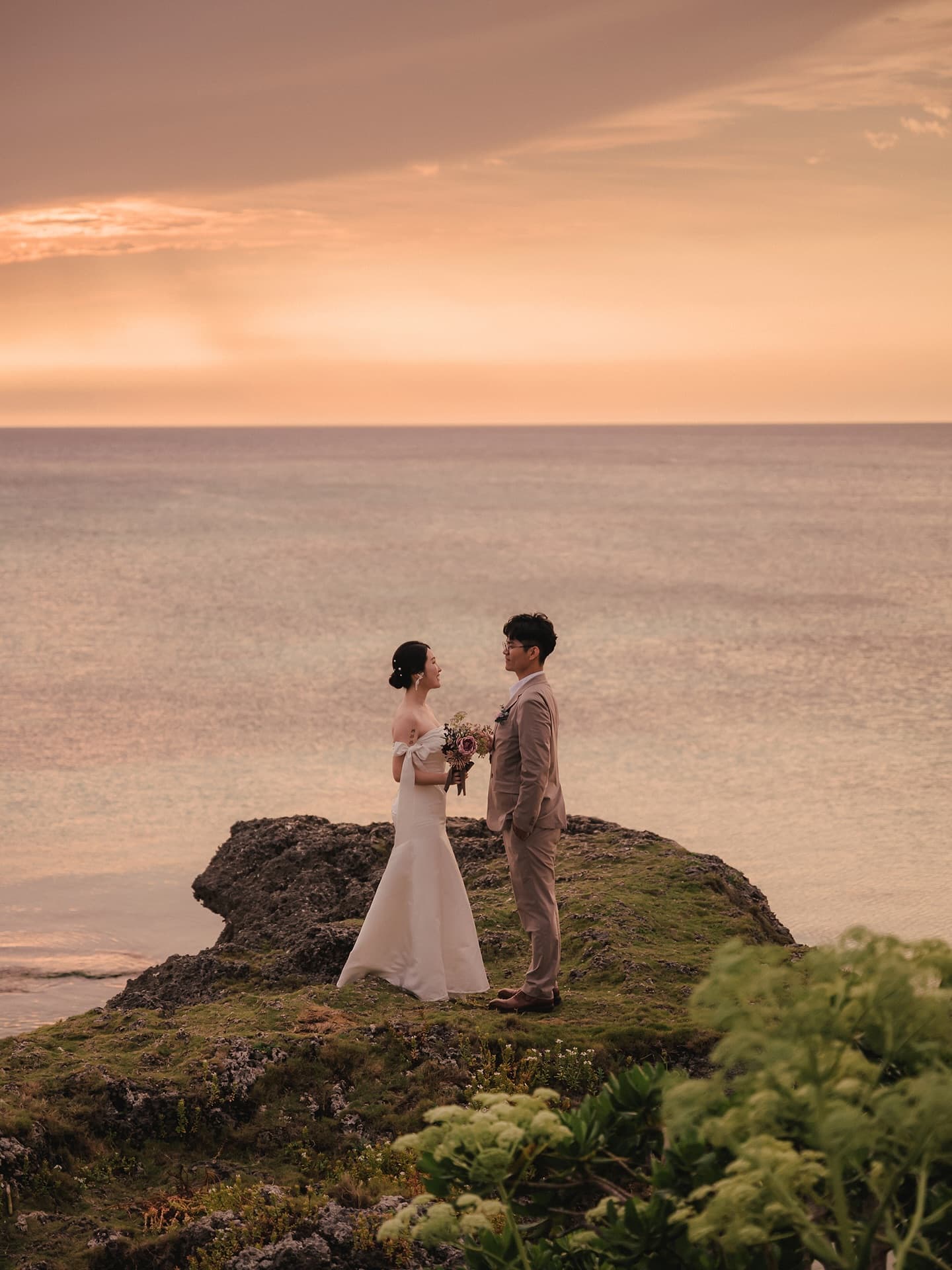 Couple facing each other on a rock under a pastel Okinawa sunset