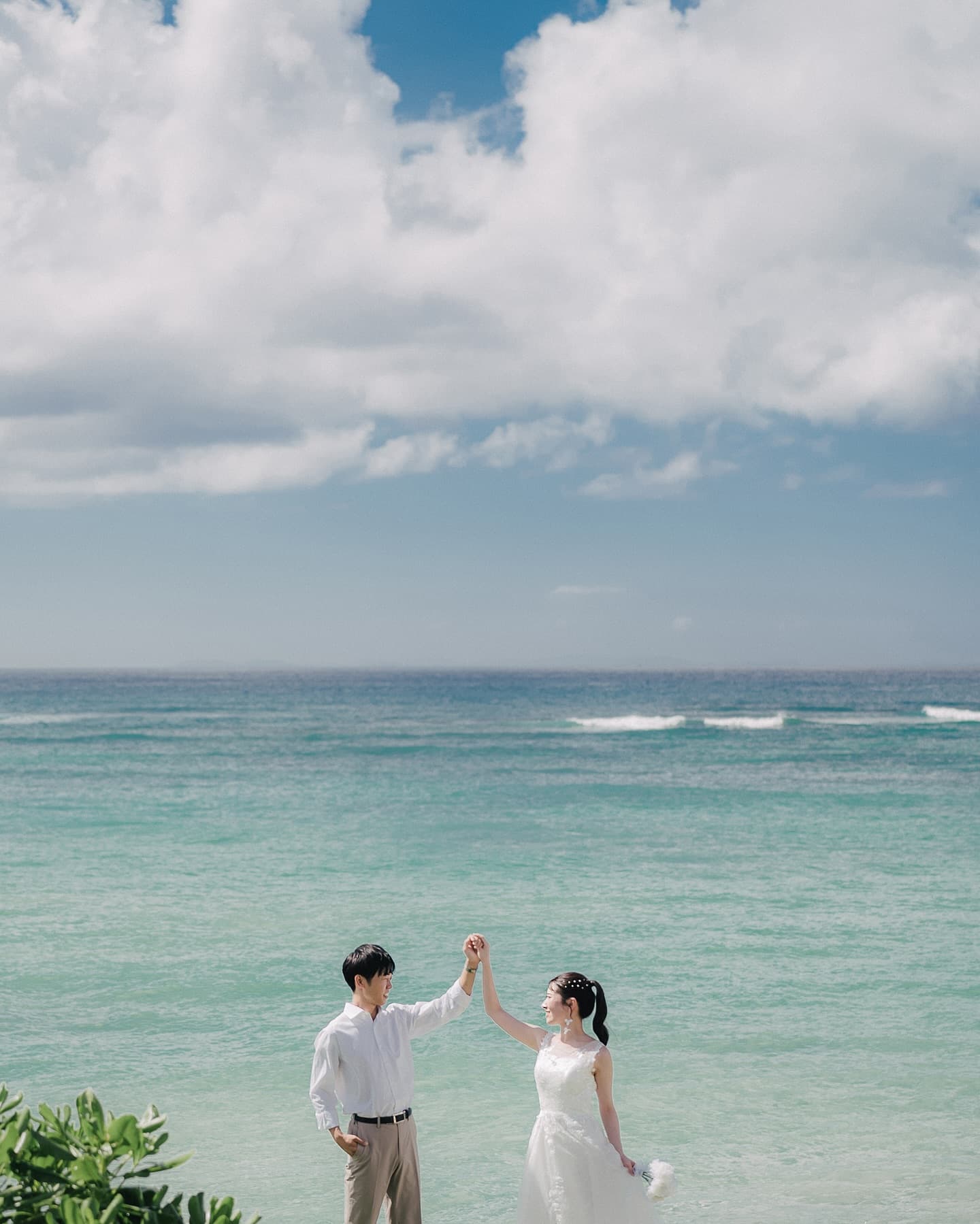 エメラルドの海を背につないだ手を掲げる新郎新婦、広い空と白い雲、沖縄