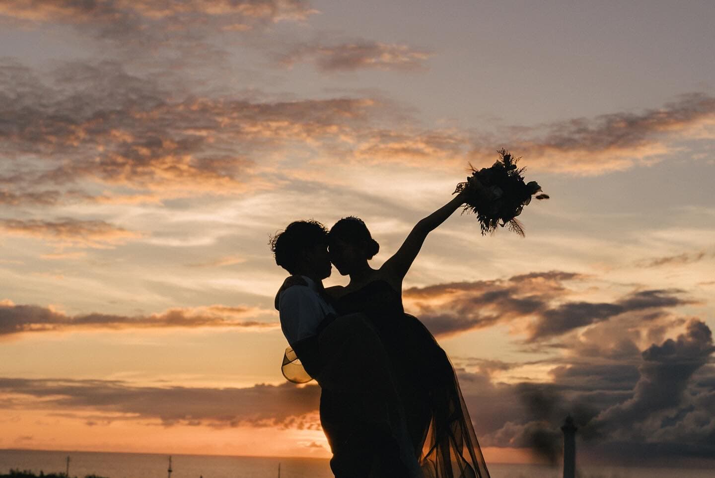 夕焼け雲の下、新婦を抱き上げブーケを掲げる二人のシルエット、沖縄