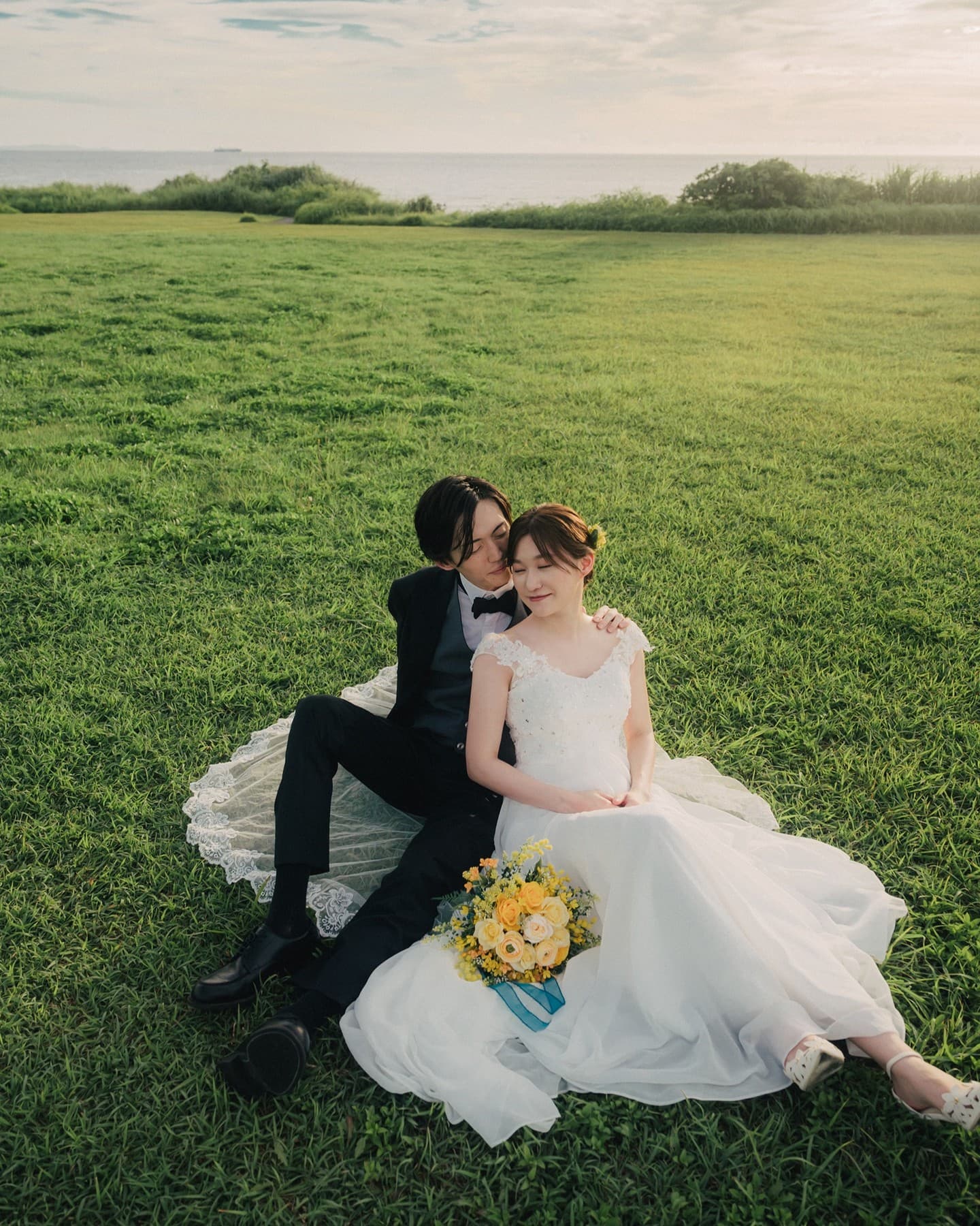 Bride and groom sit close on seaside grass in Okinawa, a yellow bouquet on her lap.