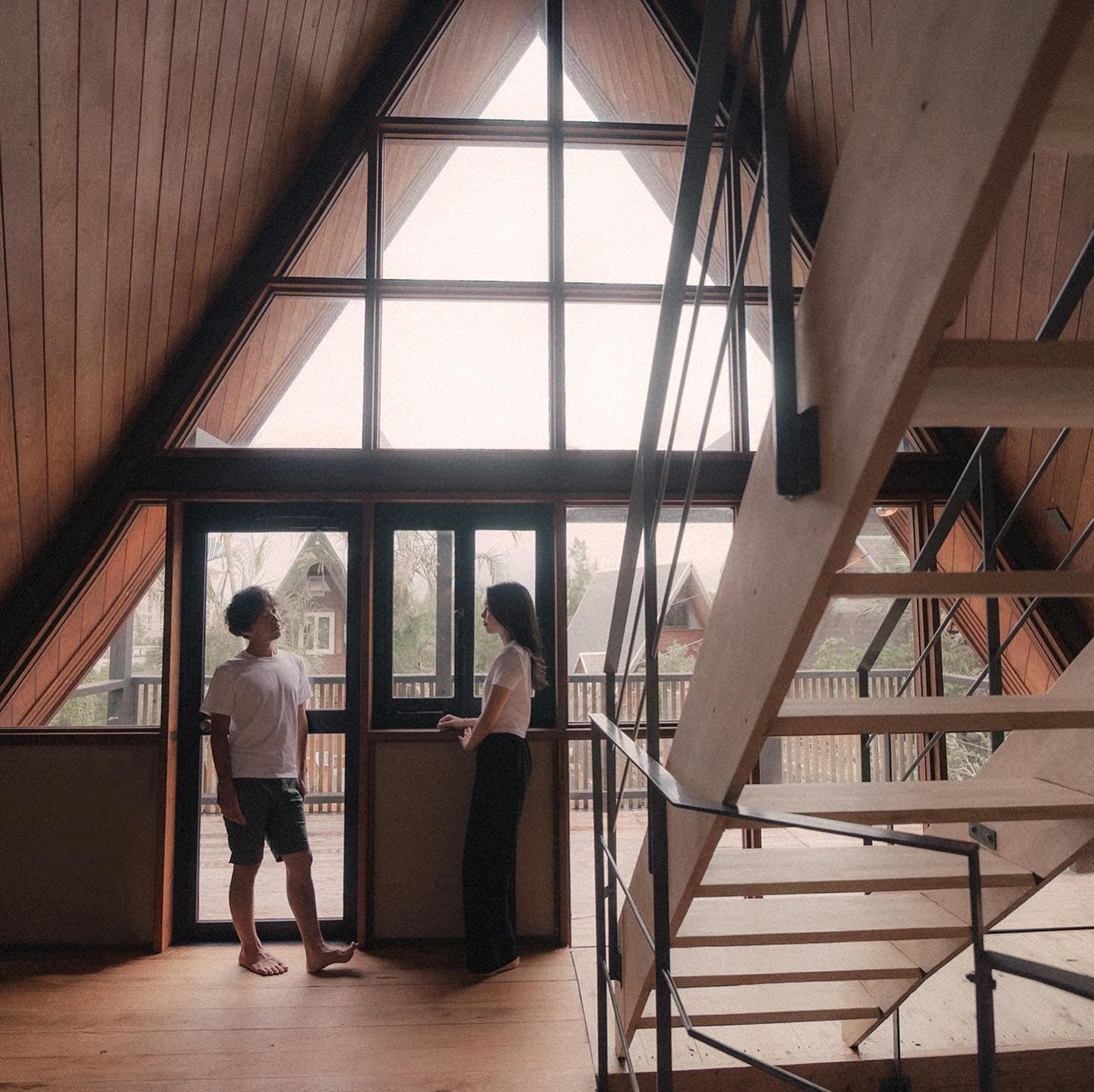 Couple in casual clothes standing by the tall triangular window of a cabin