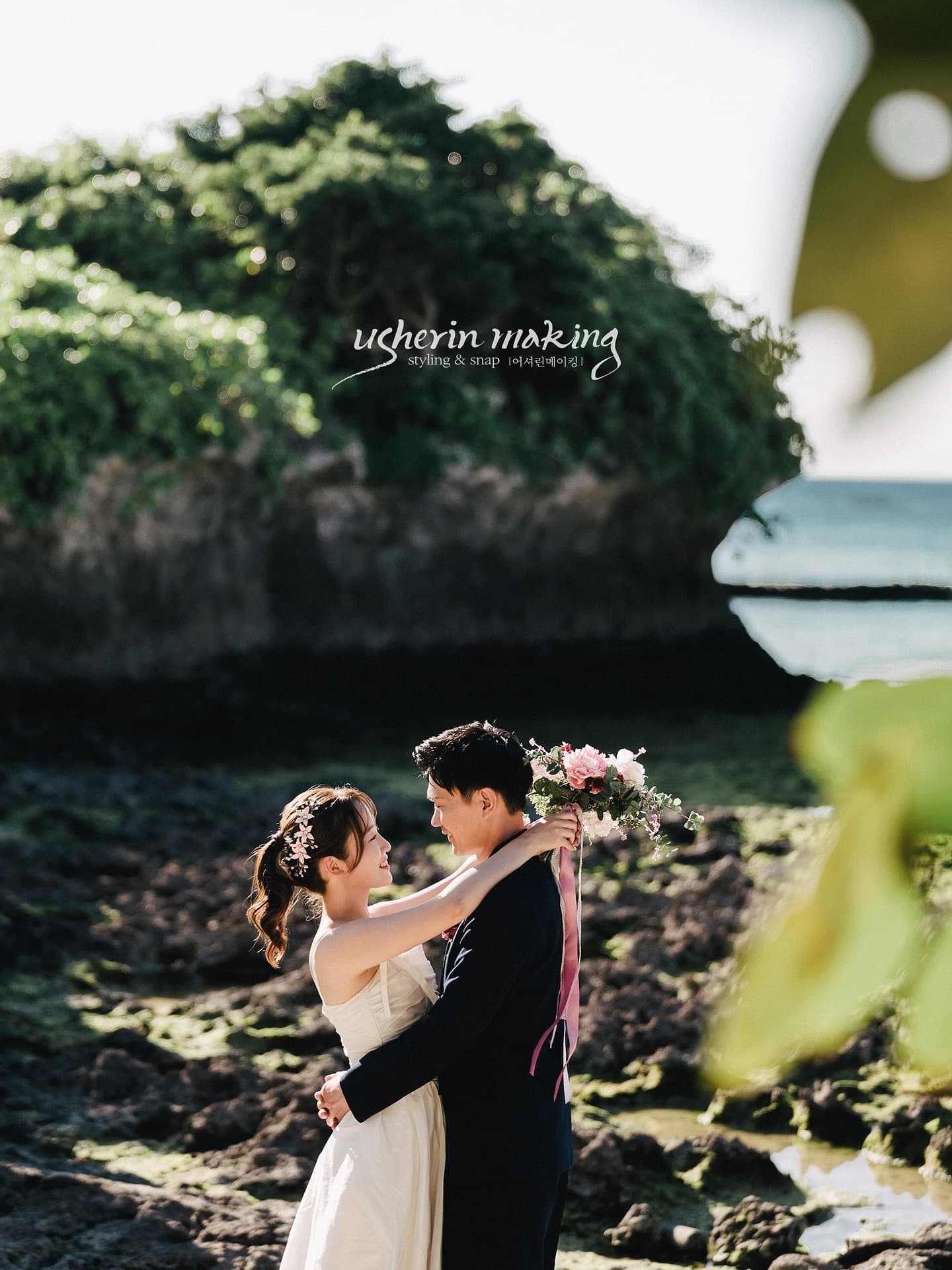 Couple embracing on a rocky shore below a green hill in Okinawa, pink bouquet with ribbons