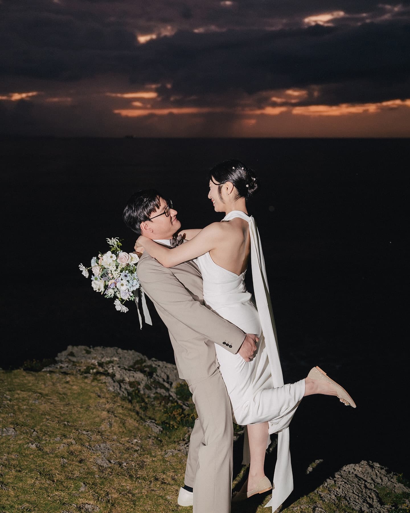 Groom lifting his bride on an Okinawa cape as the last sunset glow burns on the horizon
