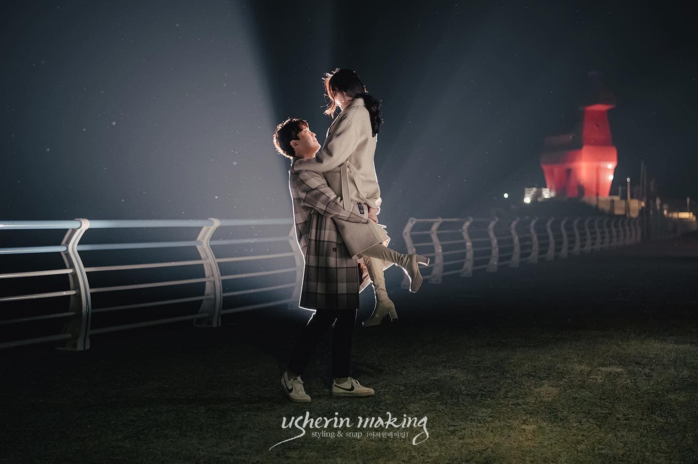 At night on Jeju Island, a man lifts his partner with a red-lit lighthouse behind them.