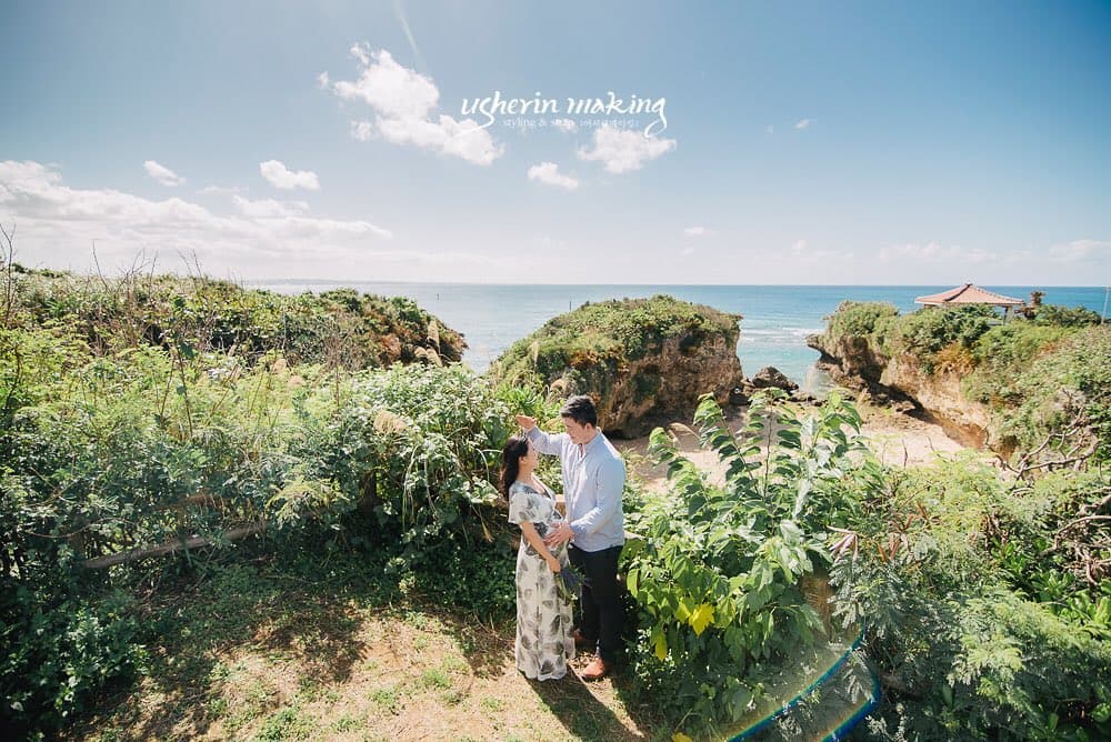 Couple standing on a lush Okinawan clifftop above the sea under a clear blue sky.