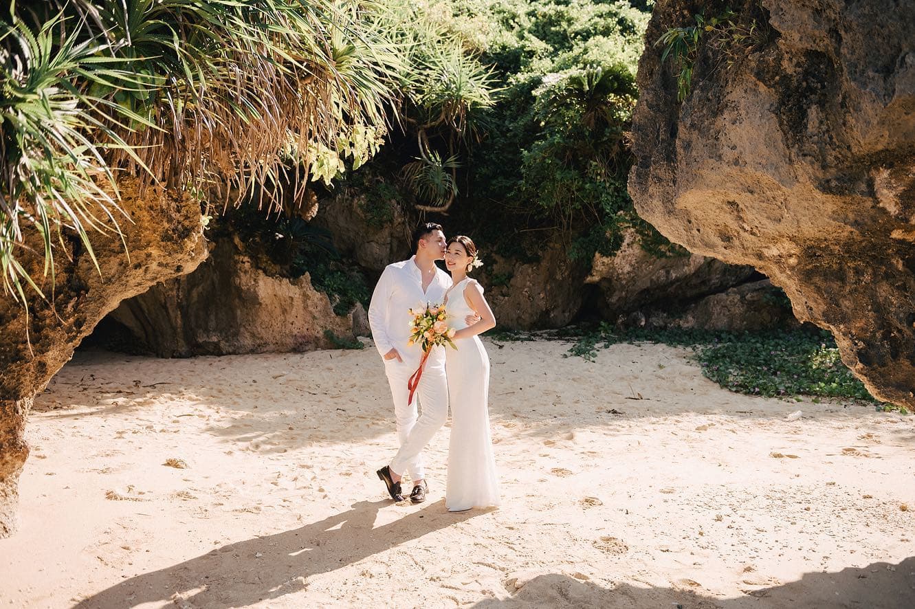 A couple in white stand together on a sandy cove framed by rocky cliffs in Okinawa