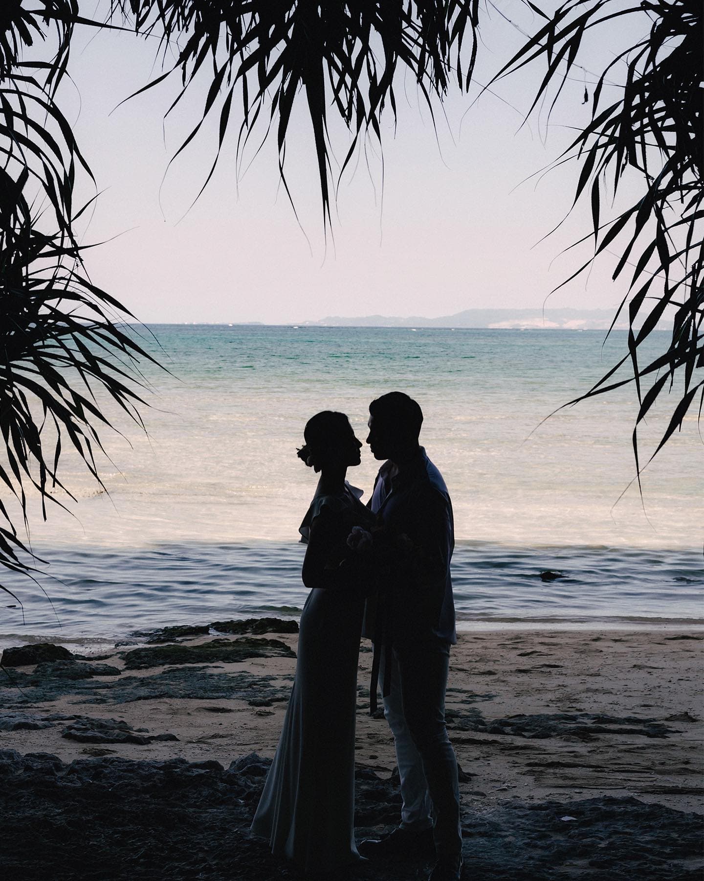 Couple silhouetted beneath palm fronds against the blue sea in Okinawa