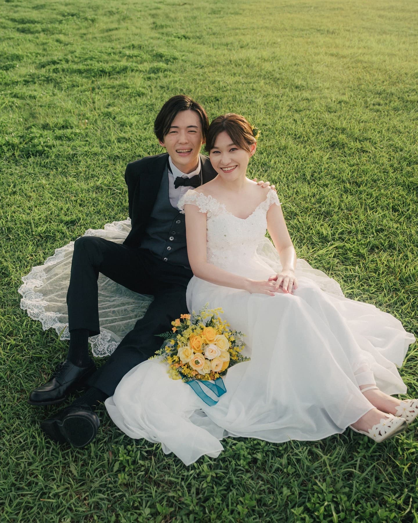 Smiling couple in wedding attire seated on the grass with a yellow rose bouquet in Okinawa.