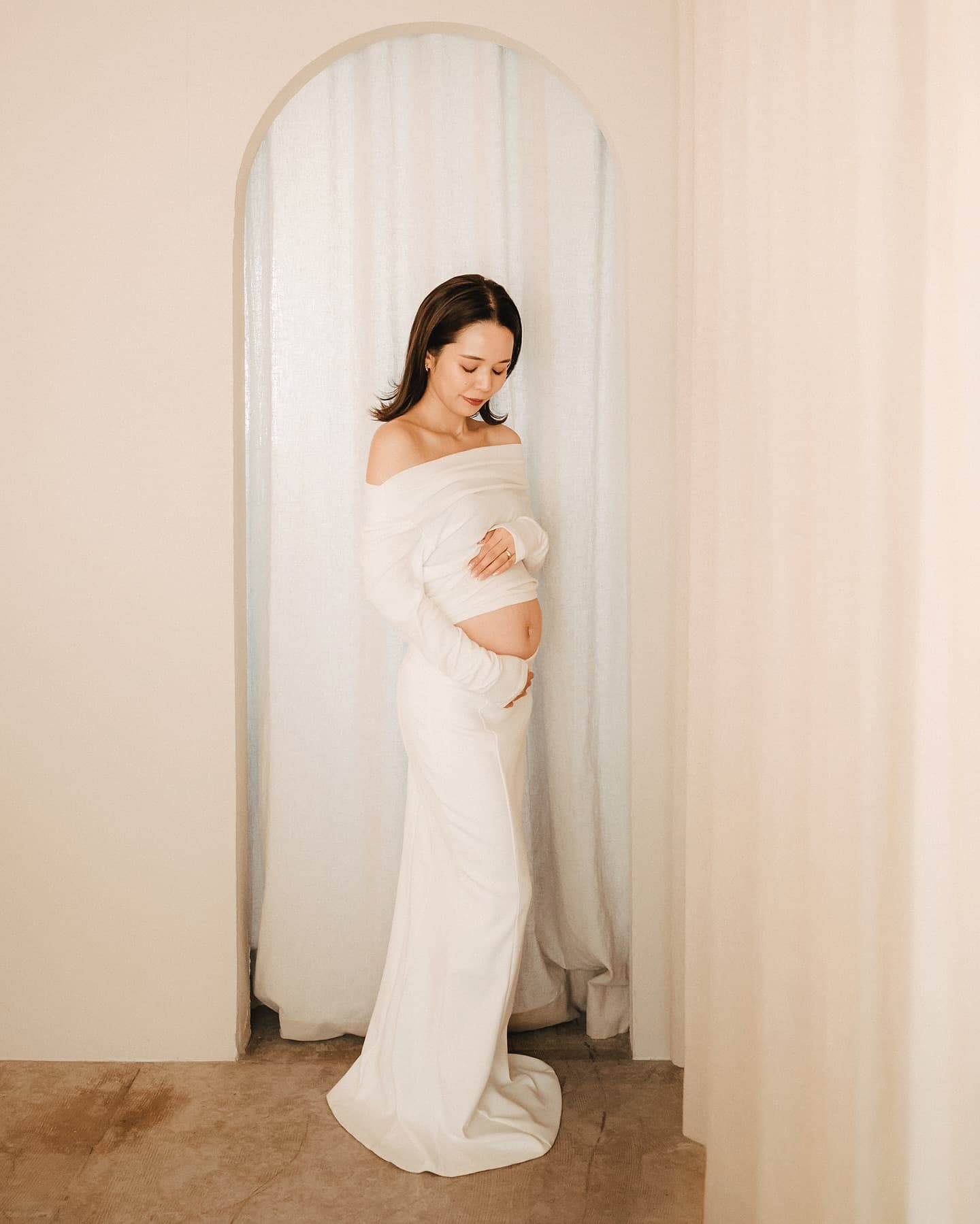 A pregnant woman in white cradles her bump by an arched curtain