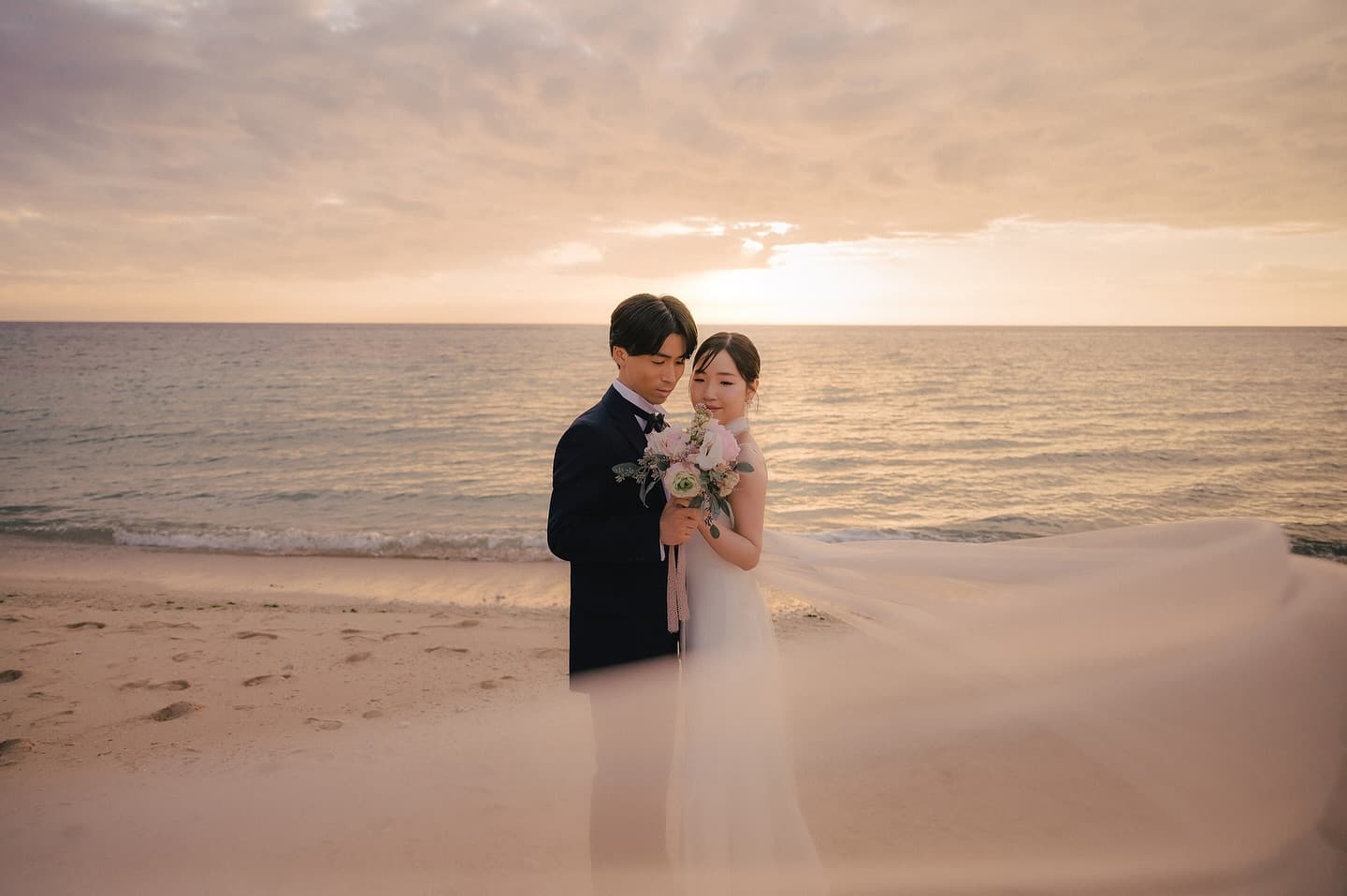 Bride and groom standing close as her long veil flows on a sunset beach in Okinawa