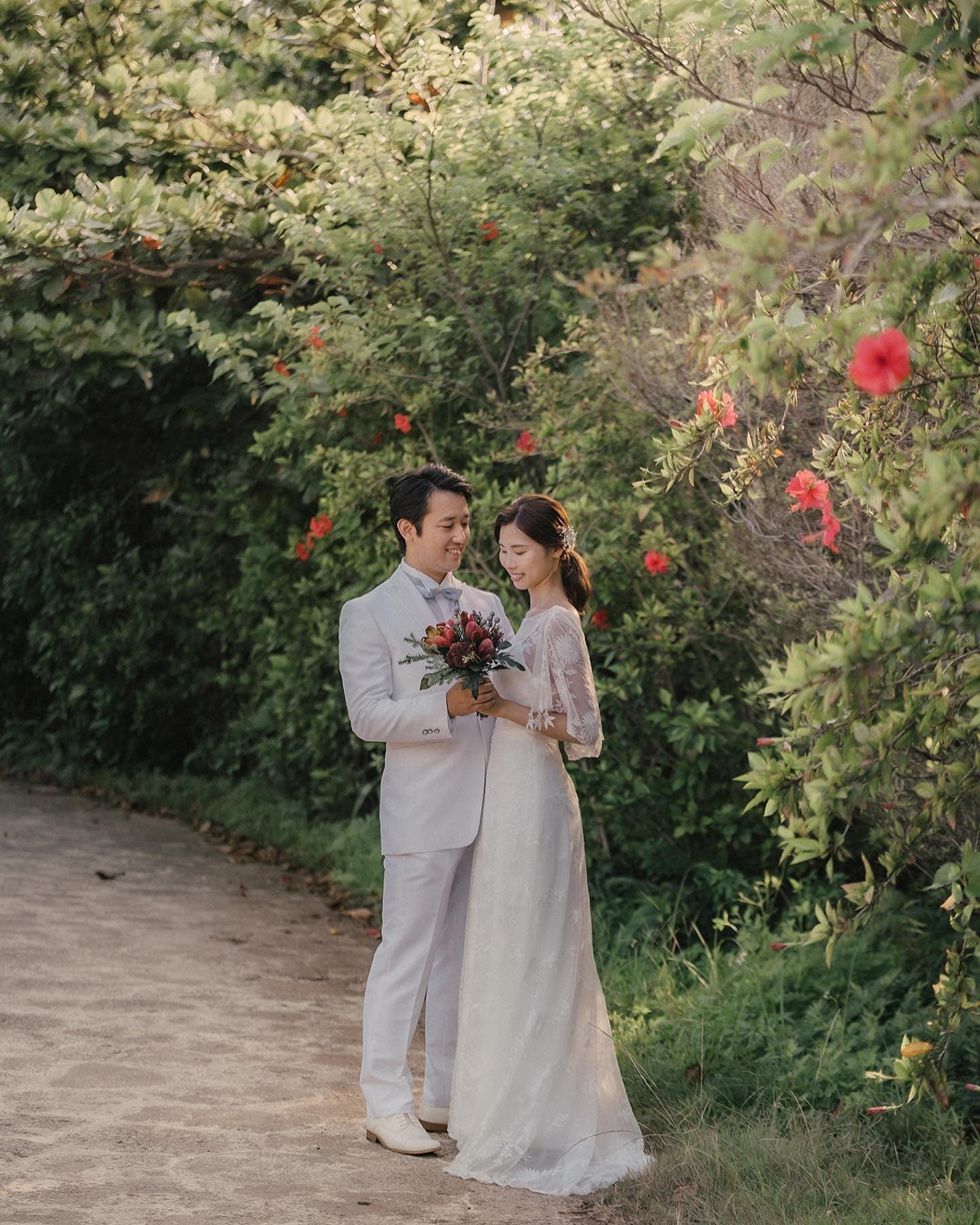 A couple in white admiring a bouquet on an Okinawa path lined with hibiscus