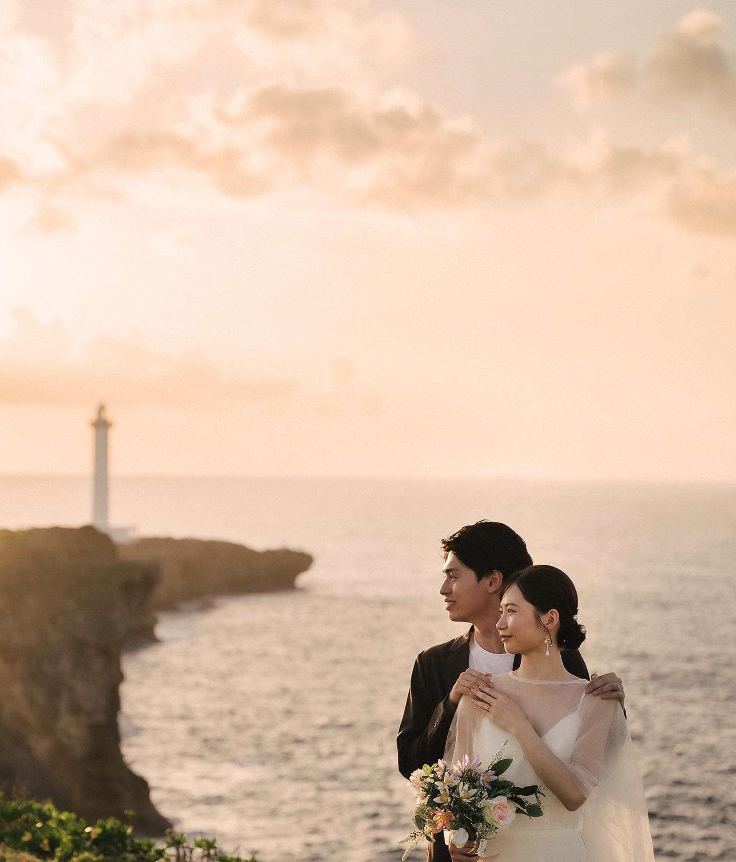 夕焼けの海を並んで眺める二人、ブーケを持つ新婦、沖縄の灯台の岬
