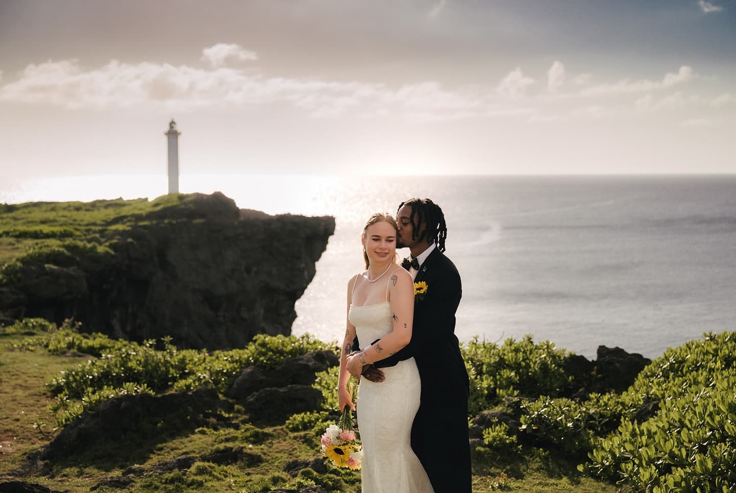 Groom embracing the bride from behind on a seaside cape with a lighthouse in Okinawa