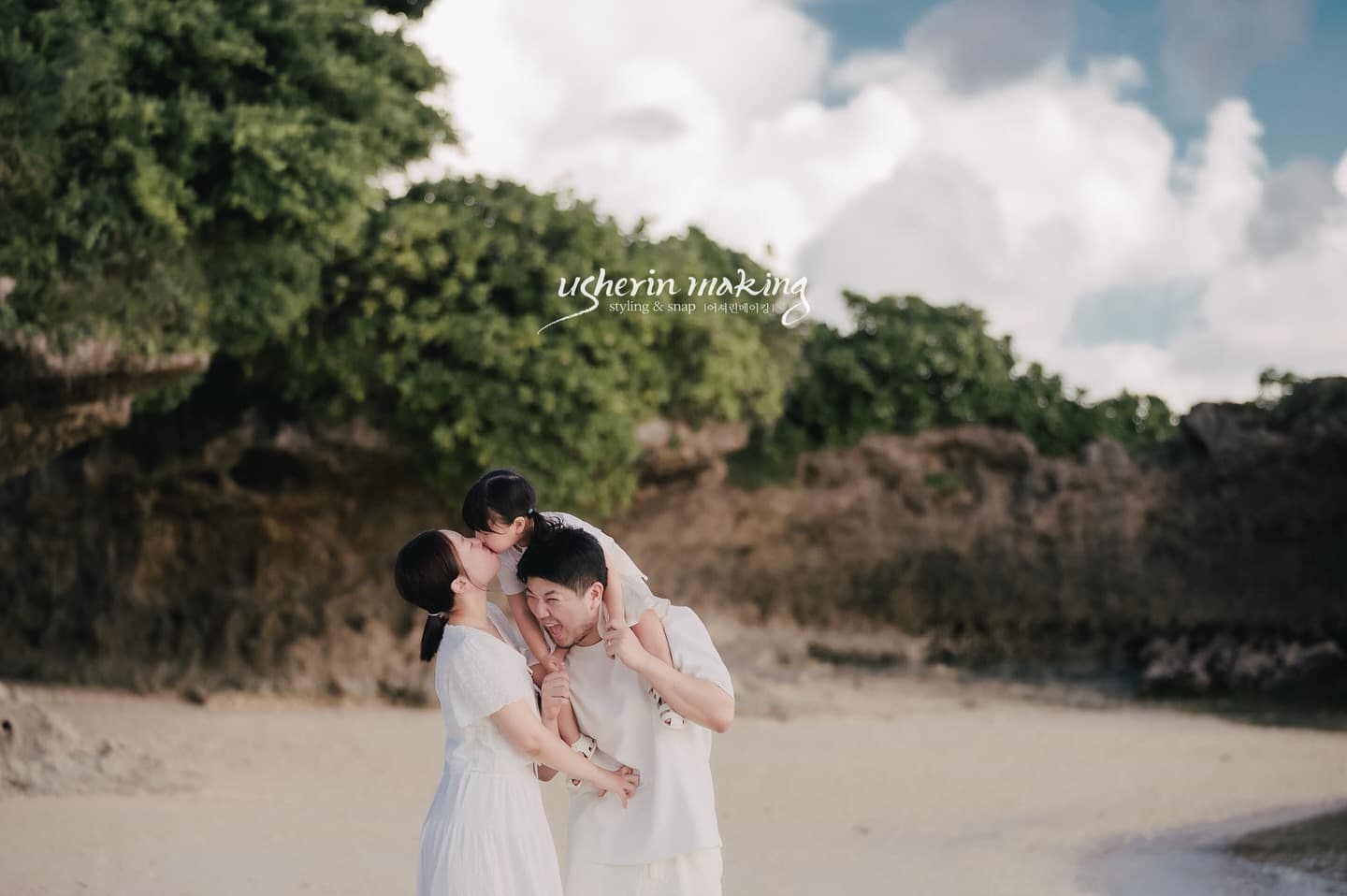 Parents in white kissing their little girl on dad's shoulders at an Okinawa beach