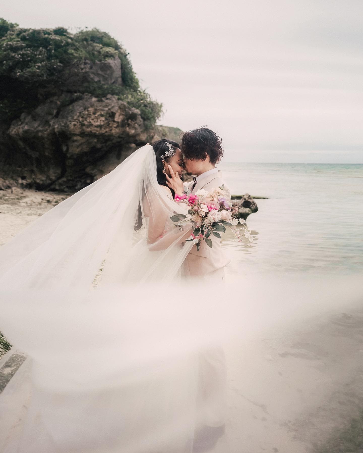 A bride's long veil sweeping across an overcast Okinawa beach as the couple embraces