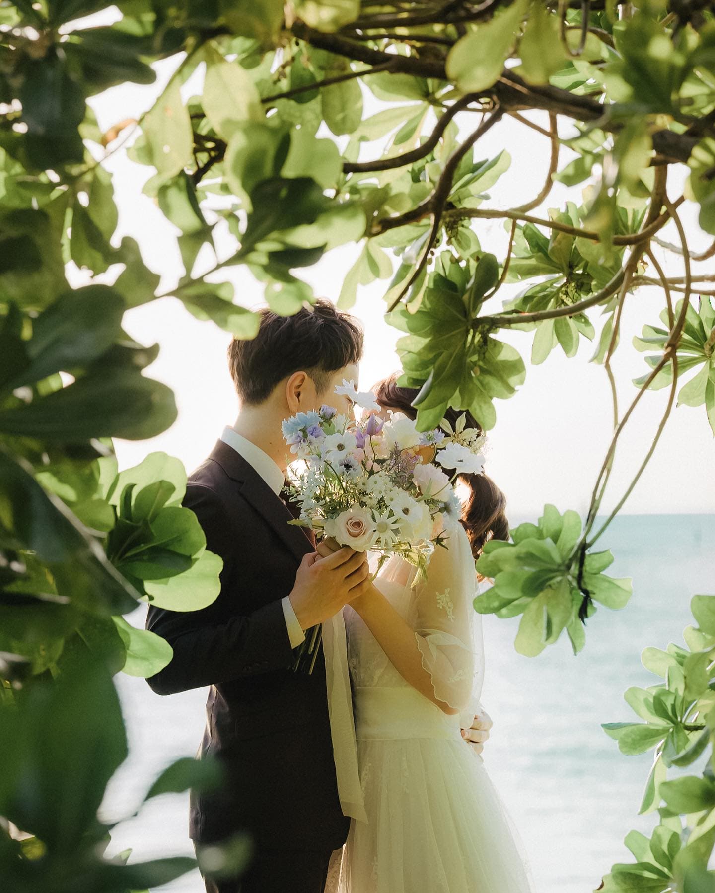 緑の葉のアーチの下、大きな花束で顔を隠す新郎新婦、沖縄の海辺