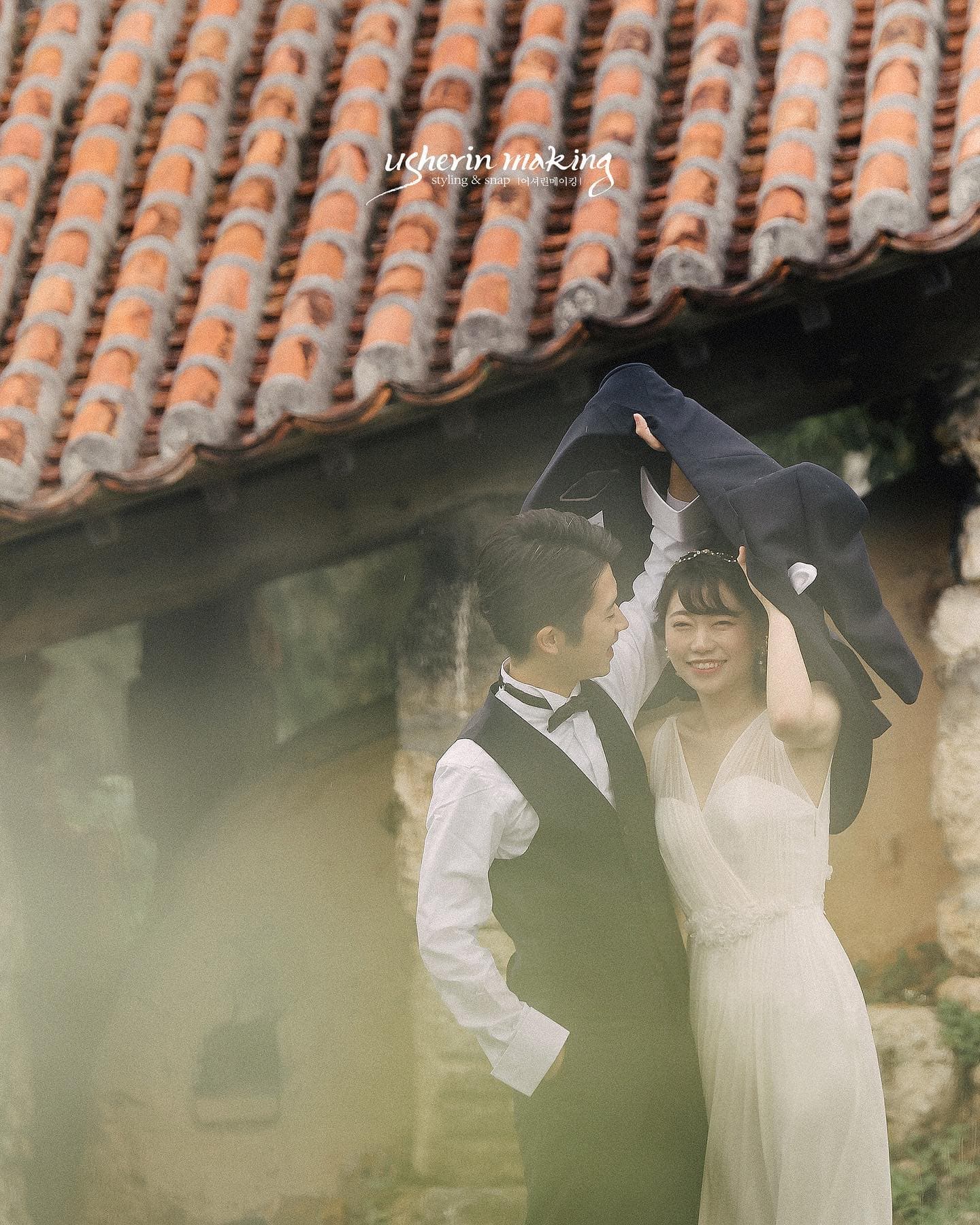 Couple in wedding attire sheltering under a jacket beneath a red-tiled roof in Okinawa