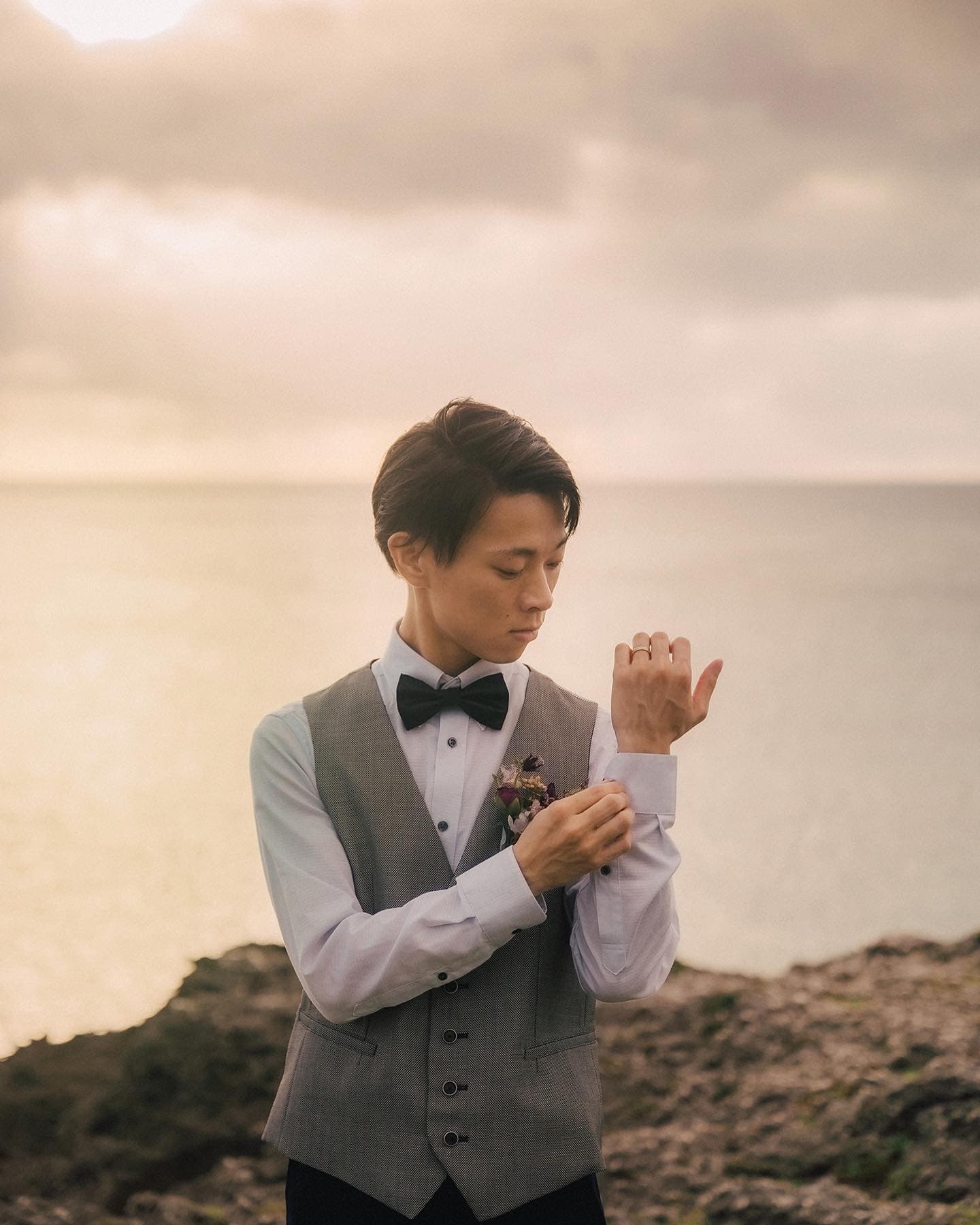 Groom in a grey vest adjusting his cuff against a moody Okinawa sea