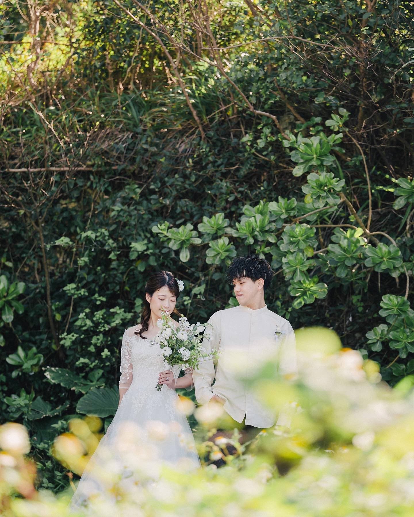 Bride in lace with a white bouquet and groom in beige amid deep Okinawa greenery