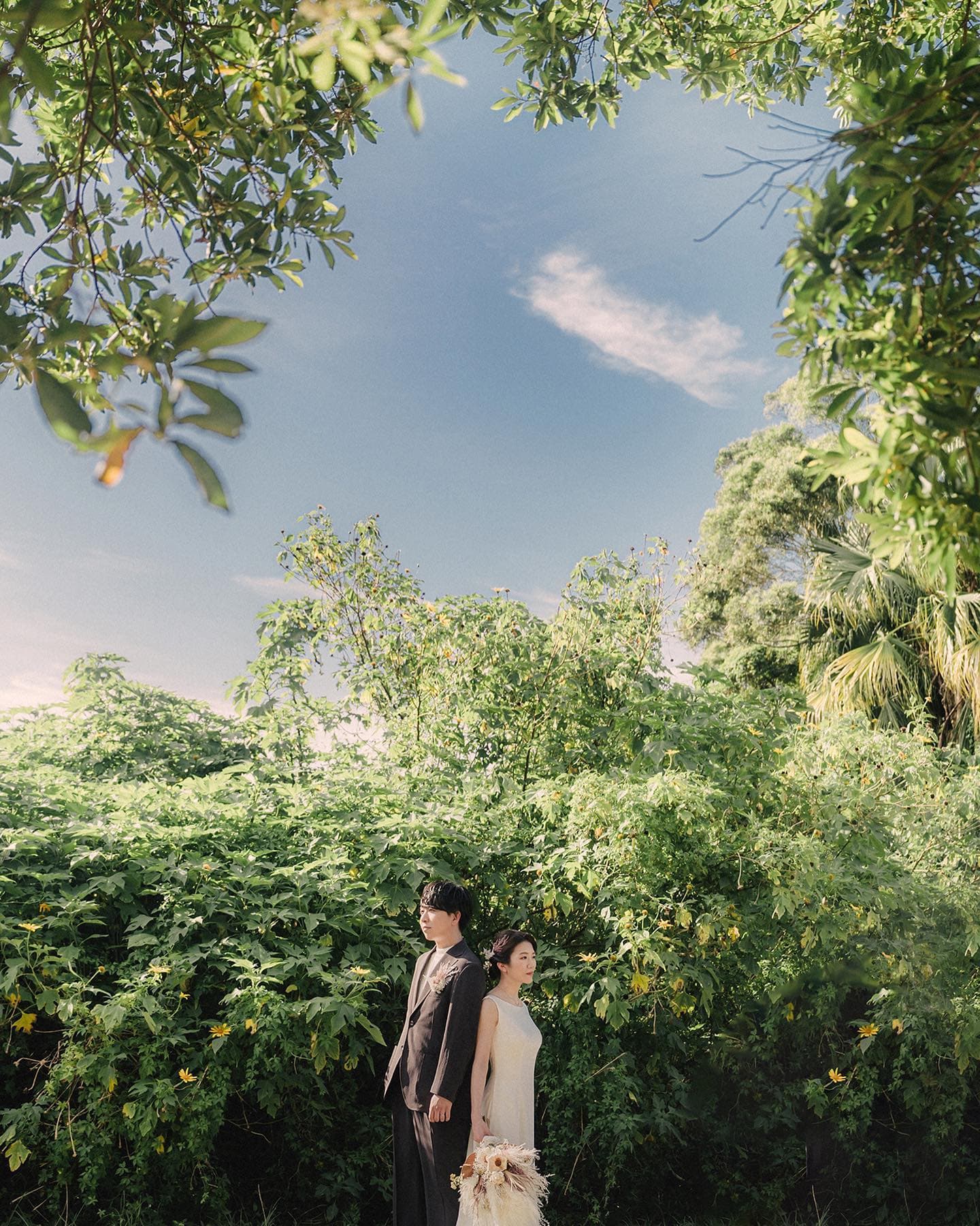 A bride and groom standing amid lush greenery under a blue Okinawa sky