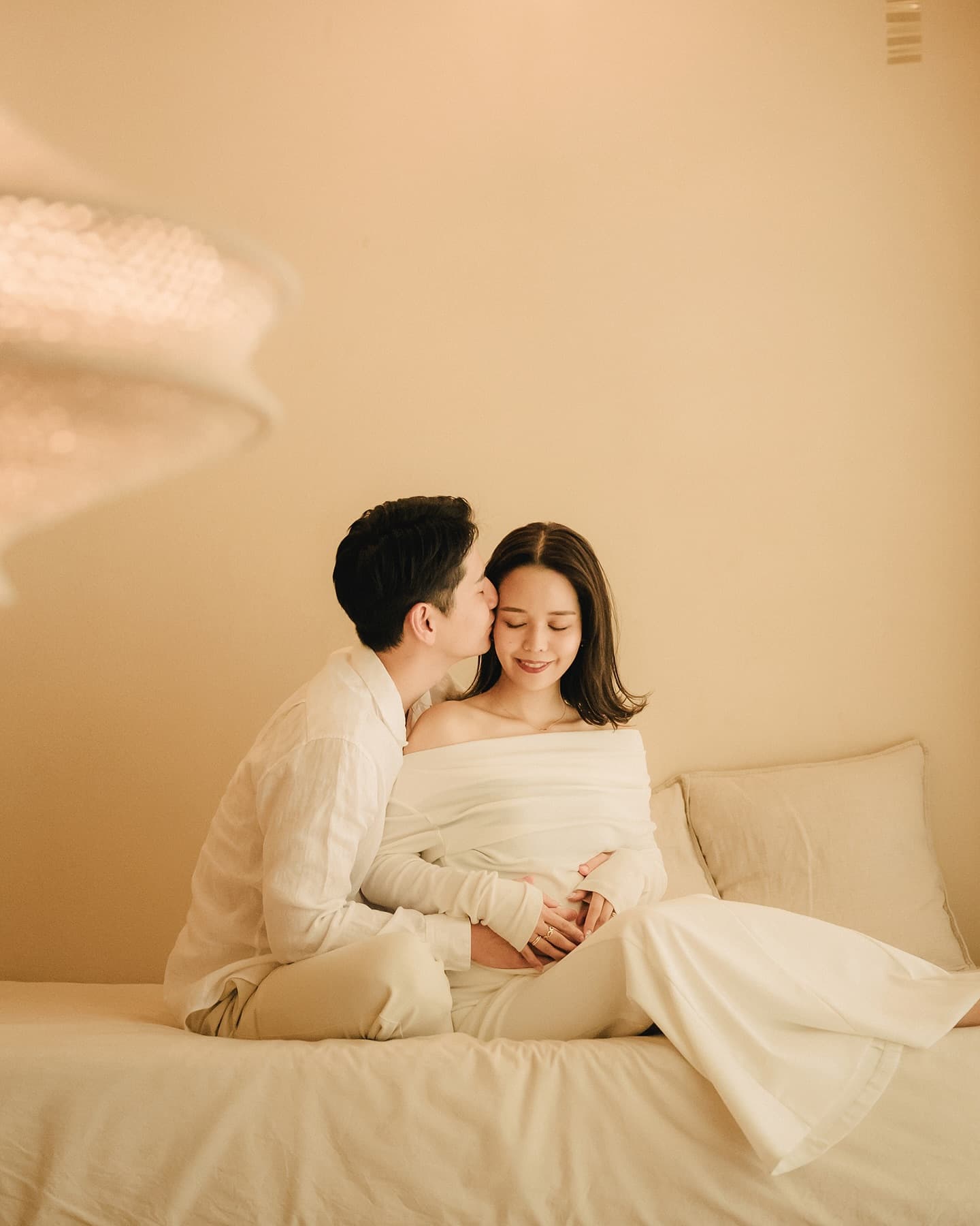 A man kisses his expecting partner's cheek on a studio bed