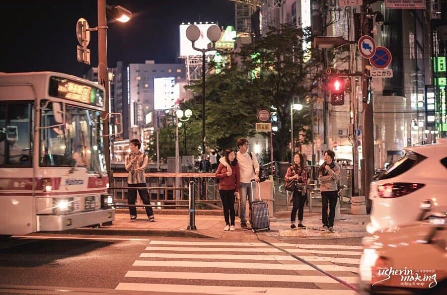 ネオンが灯る夜の街の横断歩道で、スーツケースを持って立つふたり
