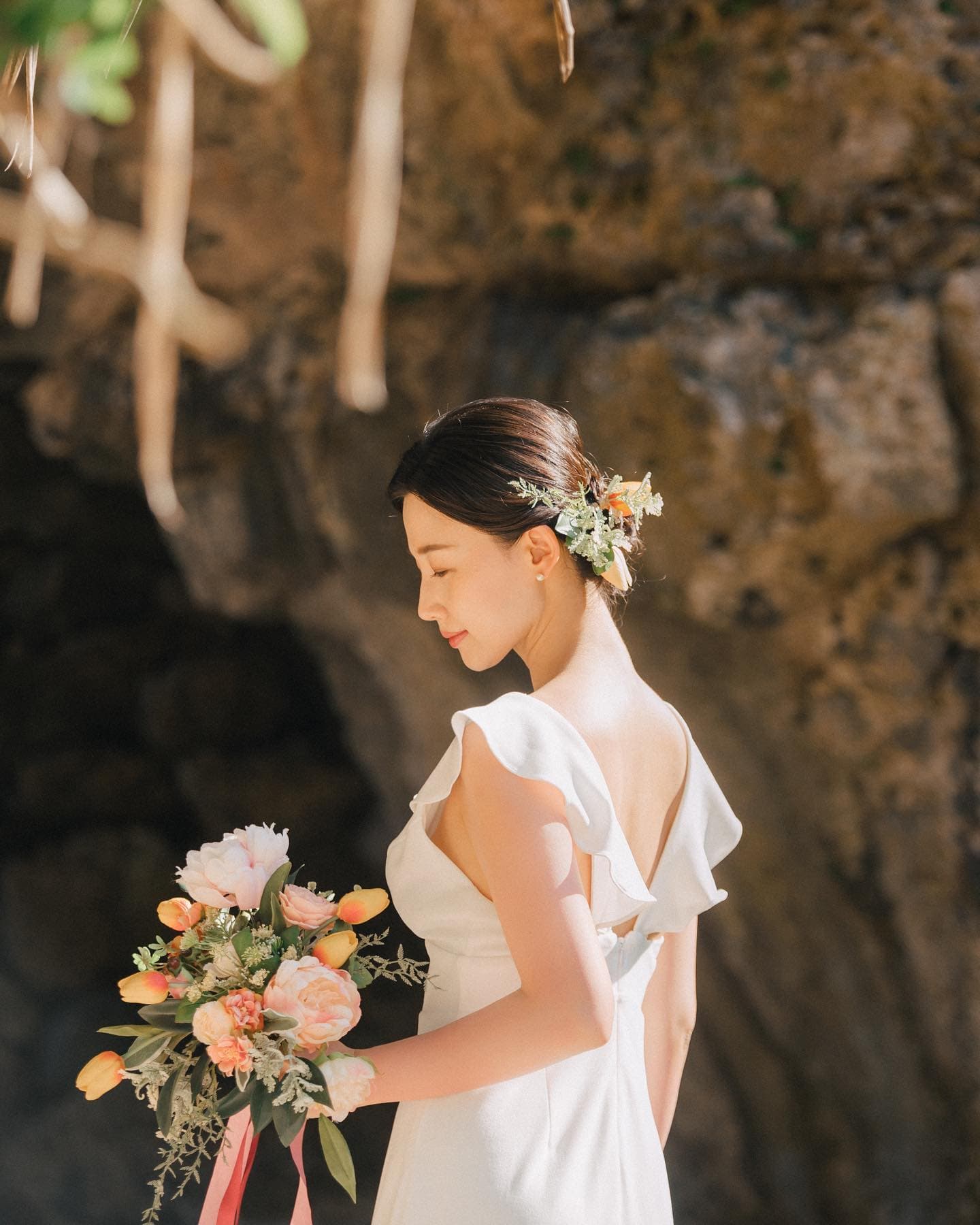 沖縄の岩場を背に、花のヘアアクセサリーとブーケを手にした花嫁の横顔。