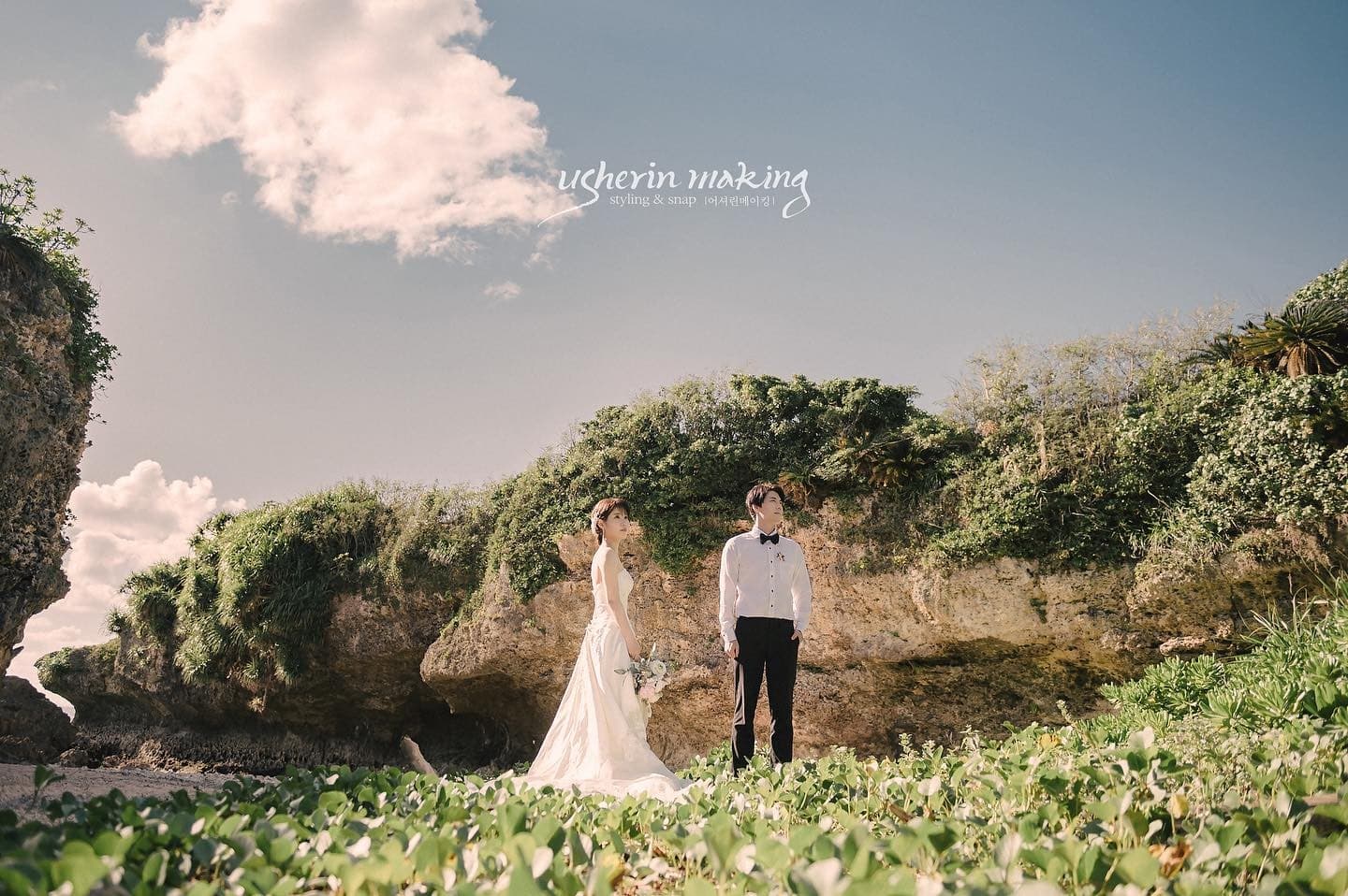 Bride and groom face each other on a lush rocky Okinawa shore under a blue sky