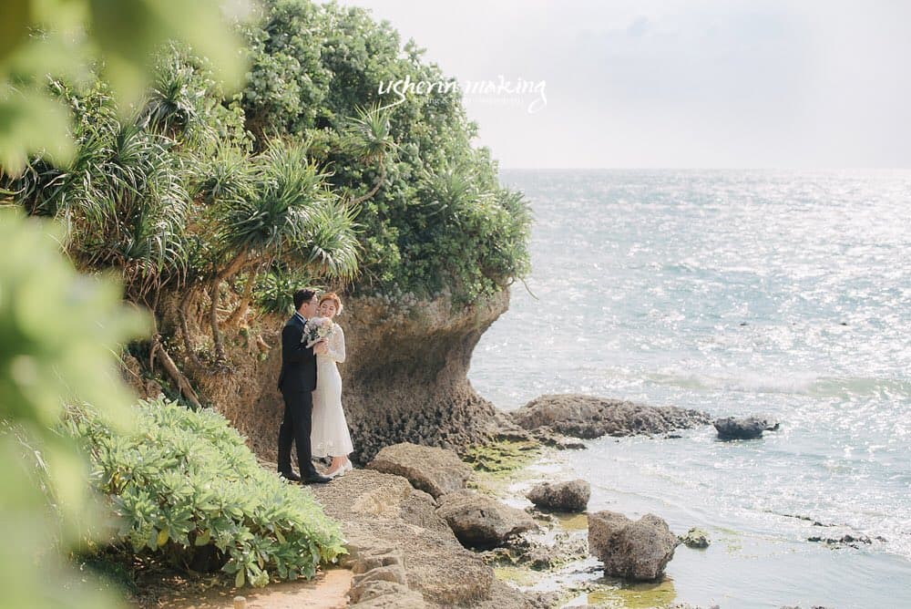 緑の岩壁ときらめく海を背に寄り添う新郎新婦、沖縄の海辺ロケーション