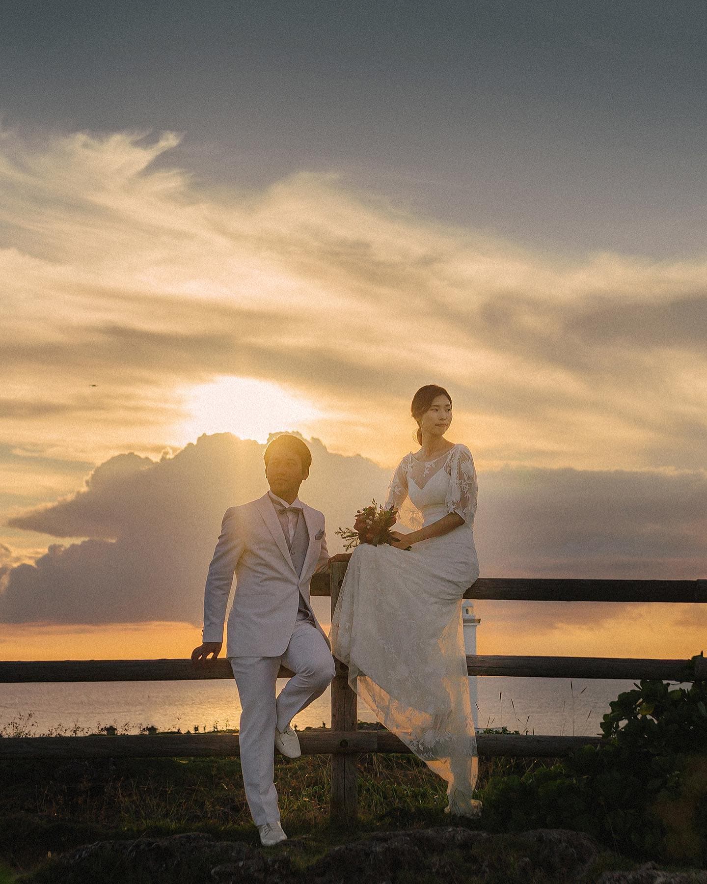 At golden hour in Okinawa, a bride sits on a wooden fence beside her groom