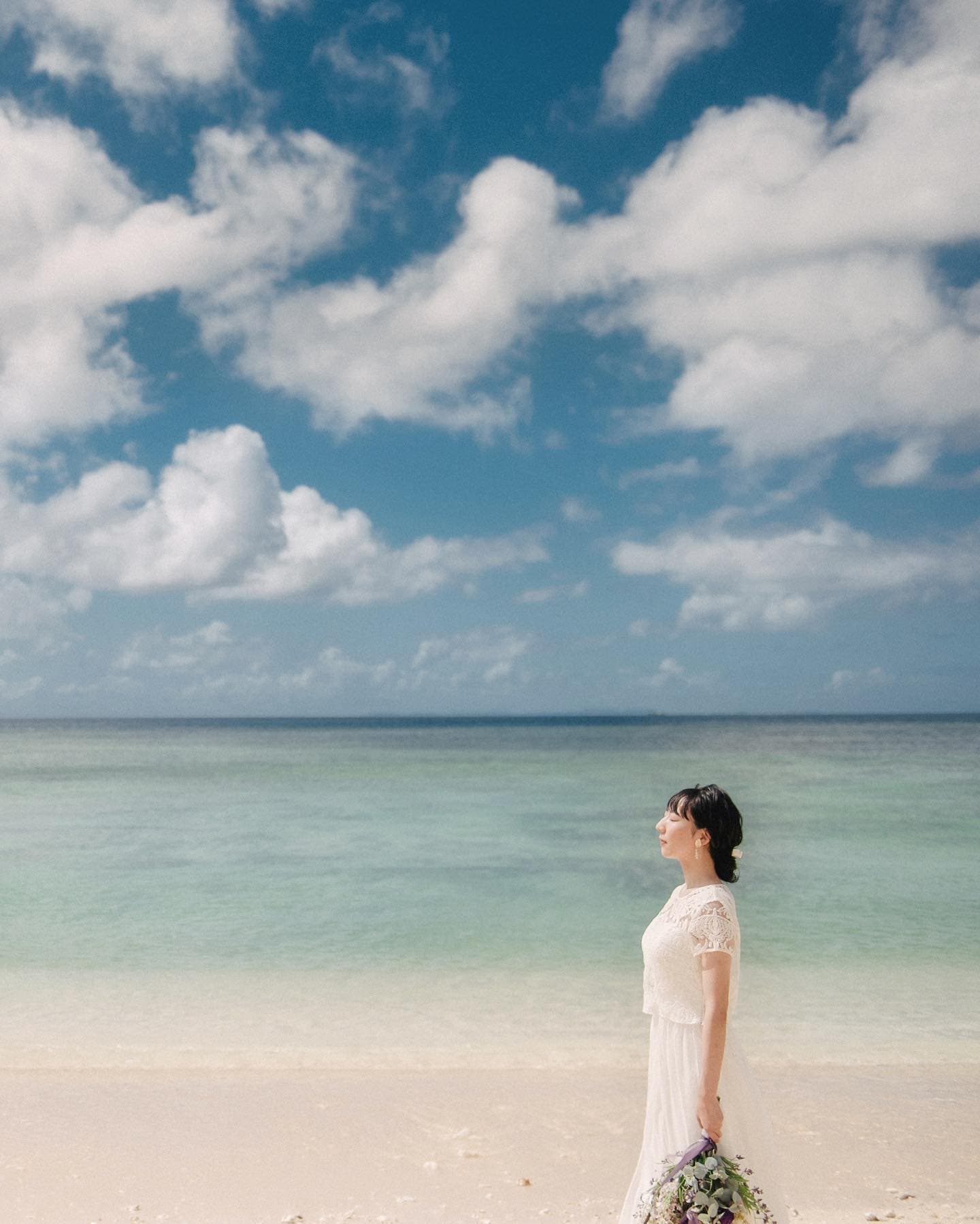 エメラルドの海を背にブーケを持って立つレースドレスの花嫁