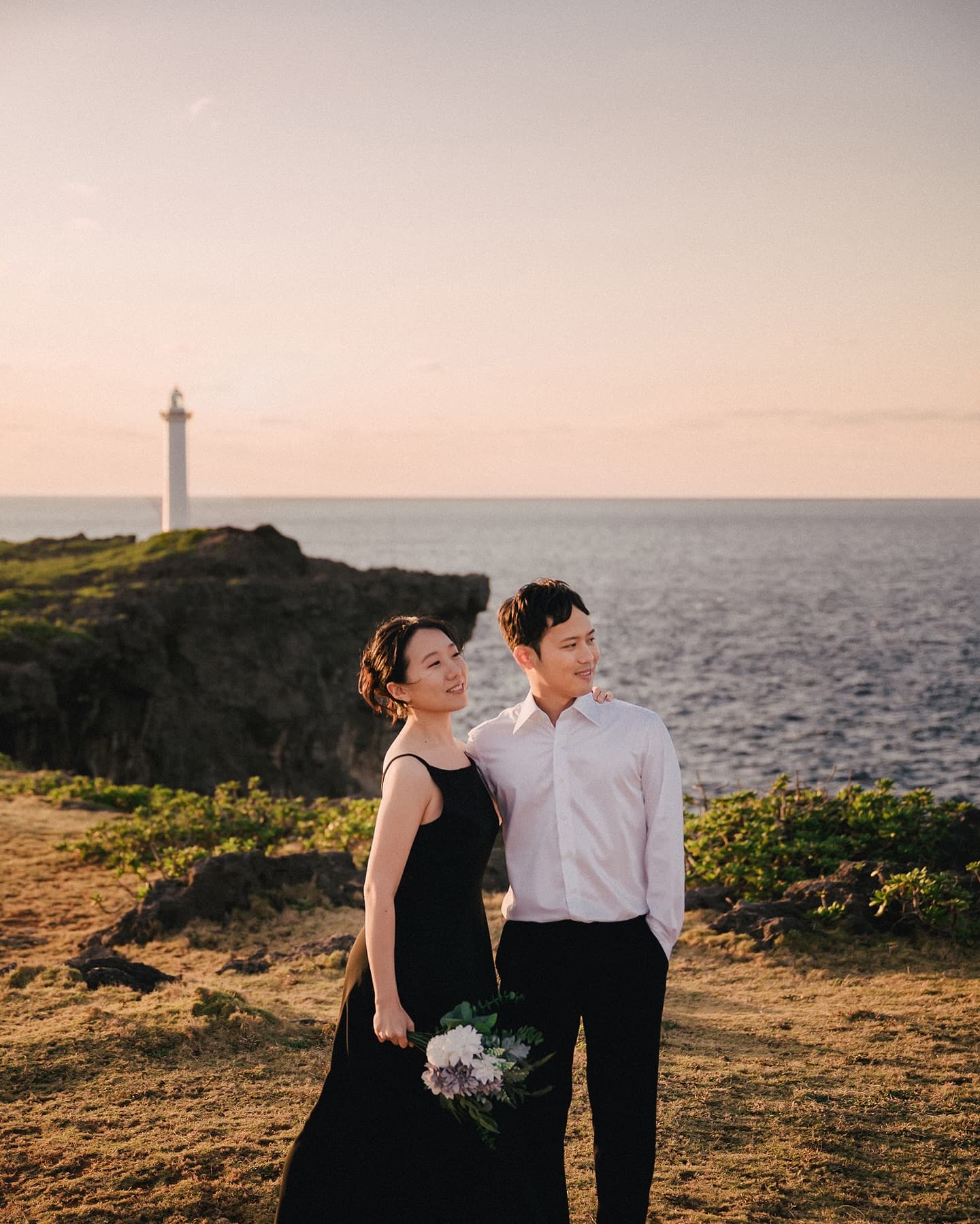 Couple embracing on a seaside cliff at dusk with a white lighthouse behind, Okinawa