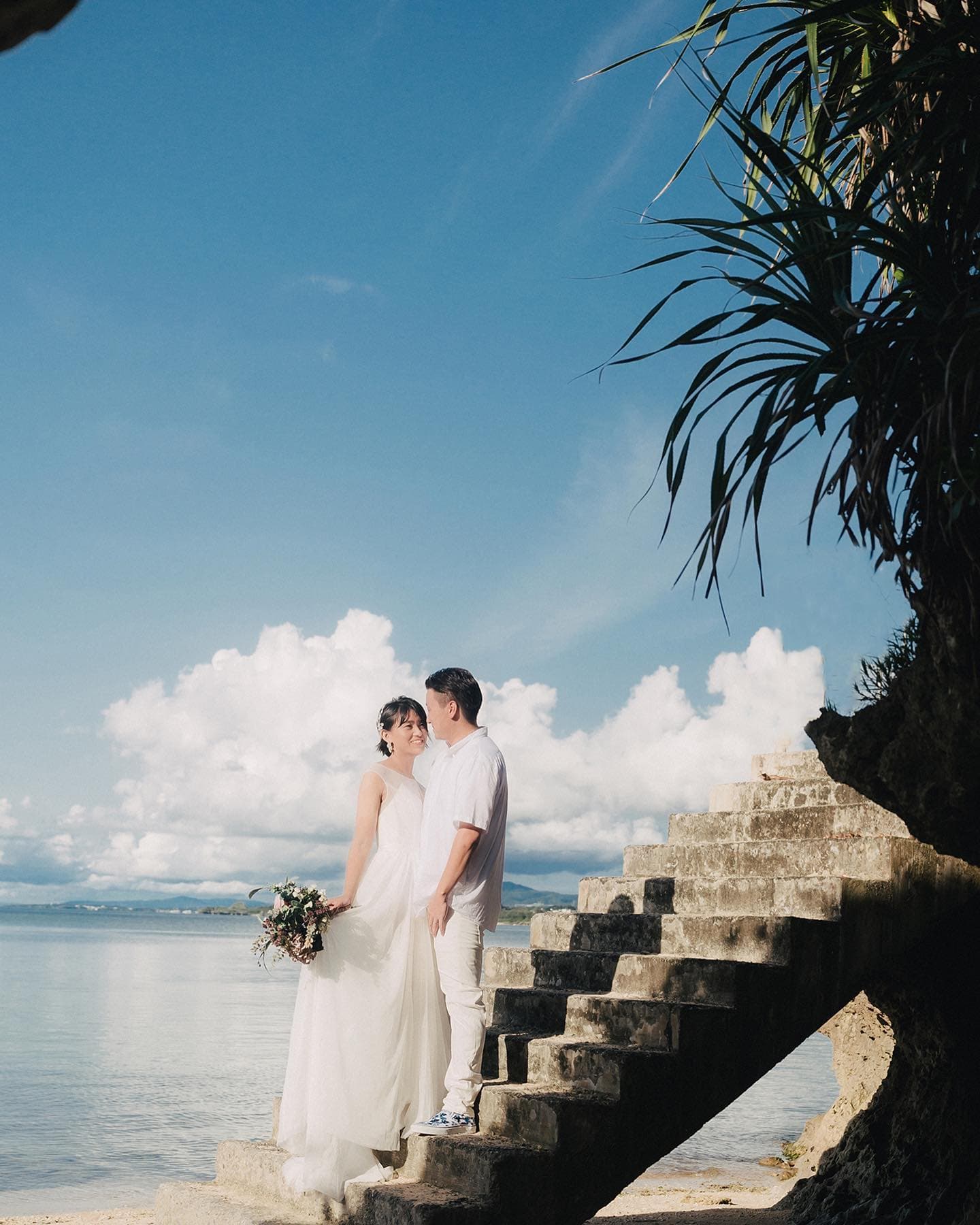 Bride and groom on stone stairs by the sea with billowing clouds in Okinawa