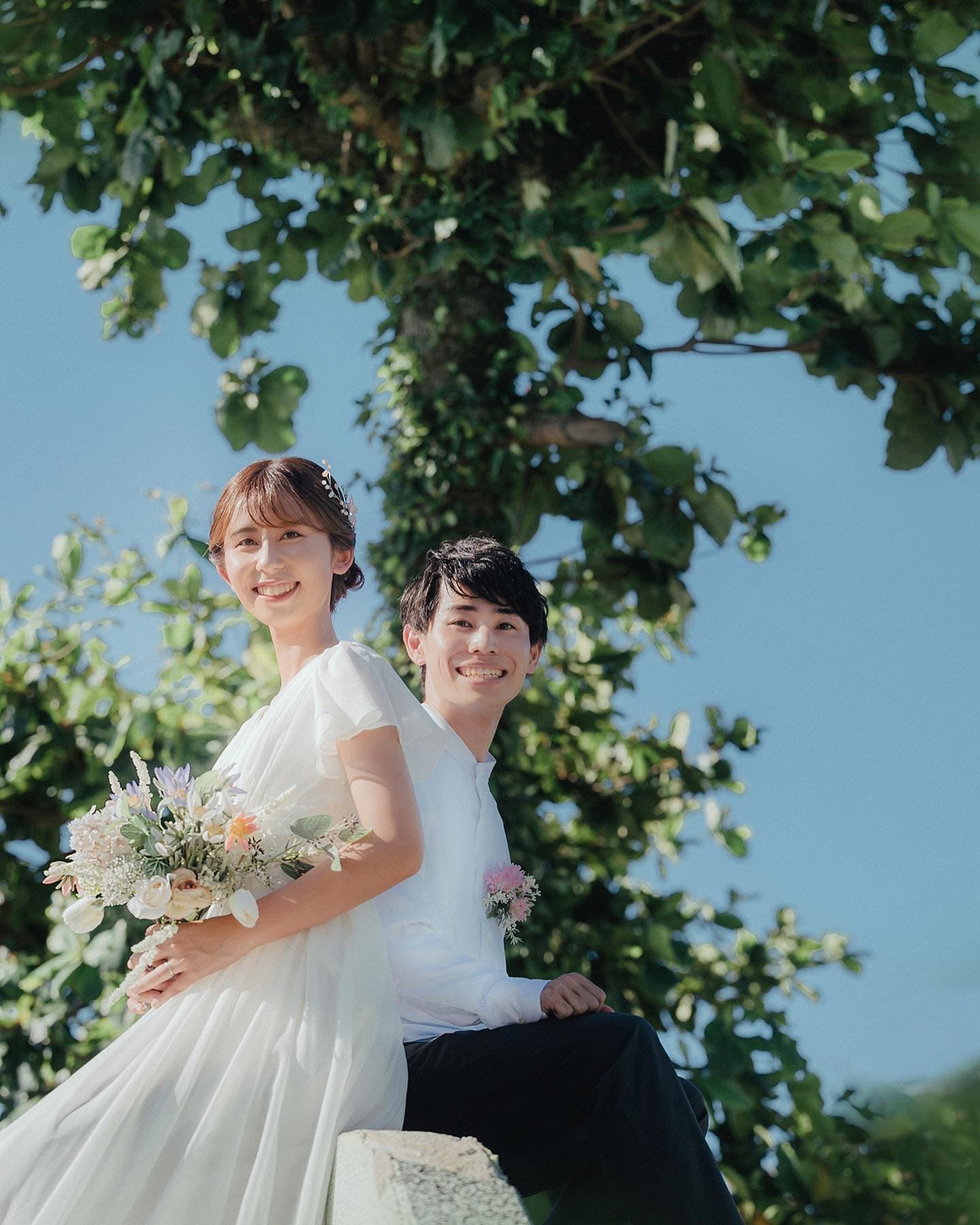 青空と緑の木を背に並んで座る花嫁と新郎