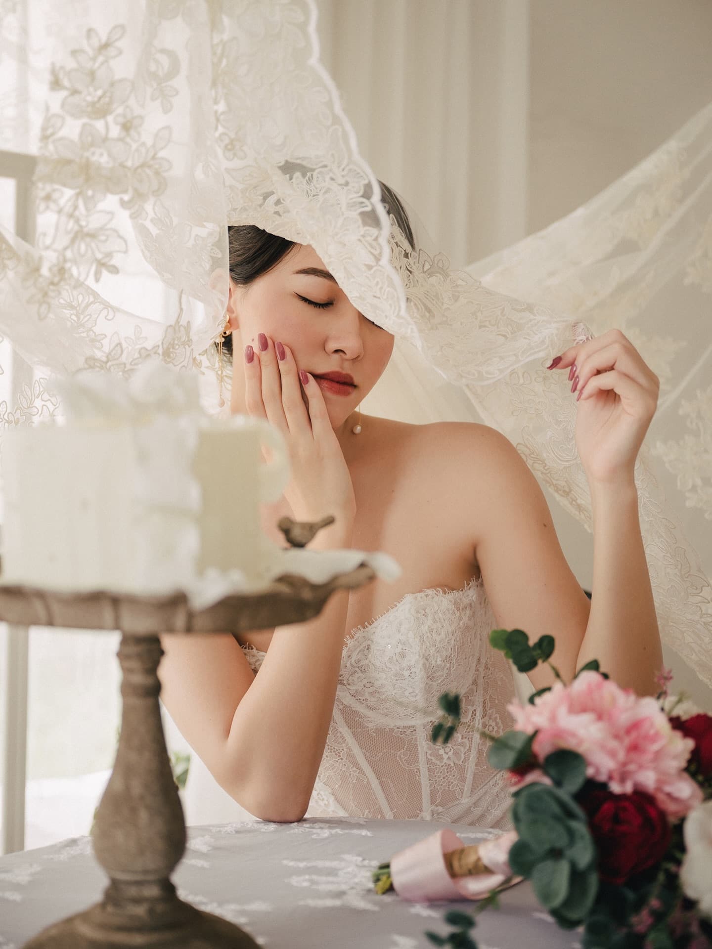 A bride closes her eyes beneath a lace veil beside a cake in the studio
