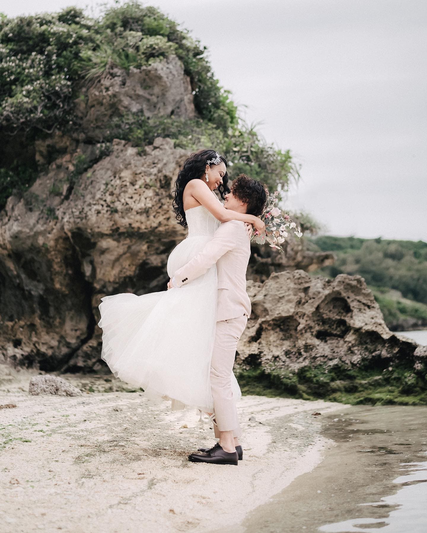 Groom lifting his bride on an Okinawa beach beneath rocky cliffs under an overcast sky