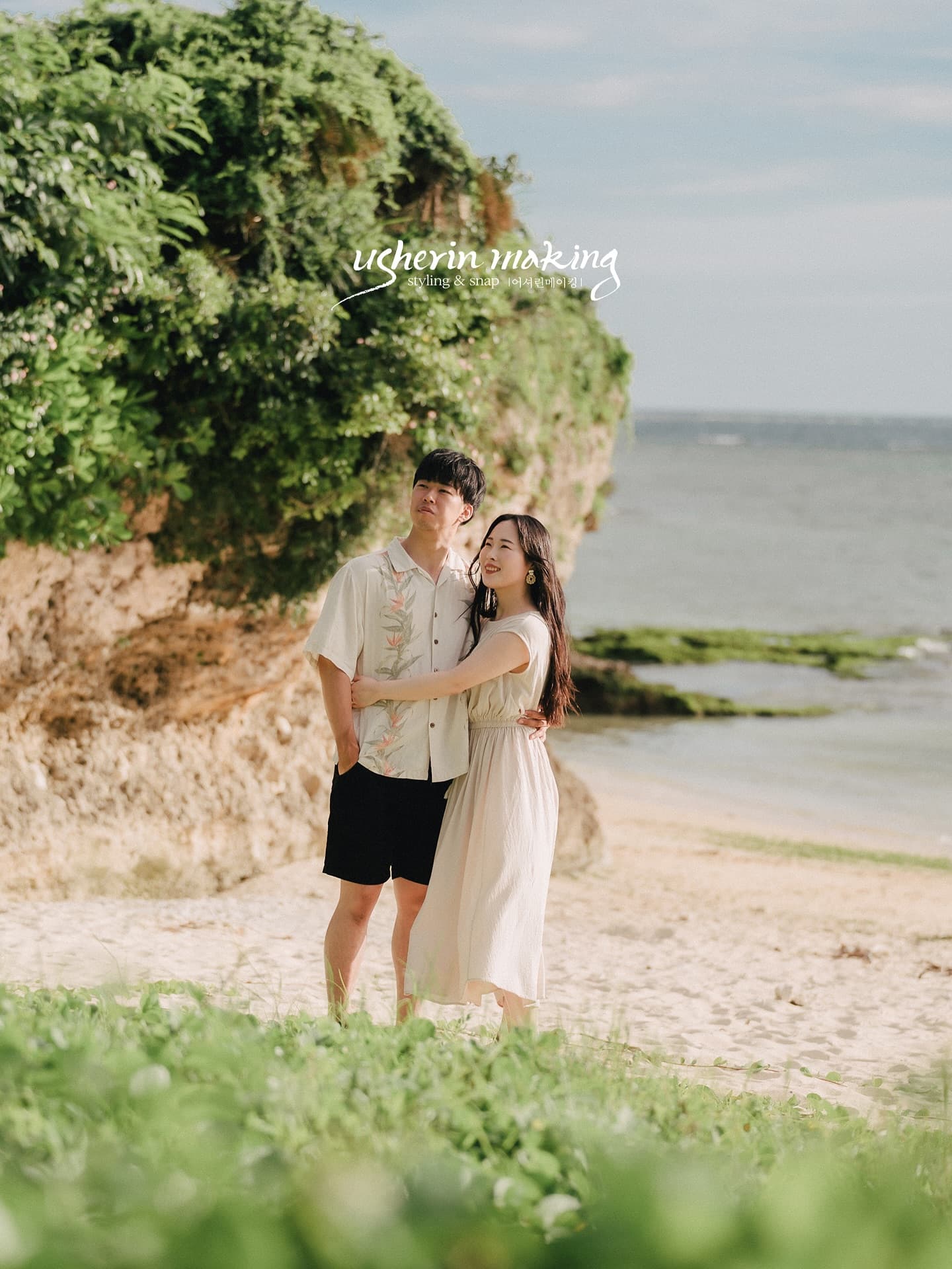 Casually dressed couple embracing on an Okinawa beach by a green-topped rock and blue sea