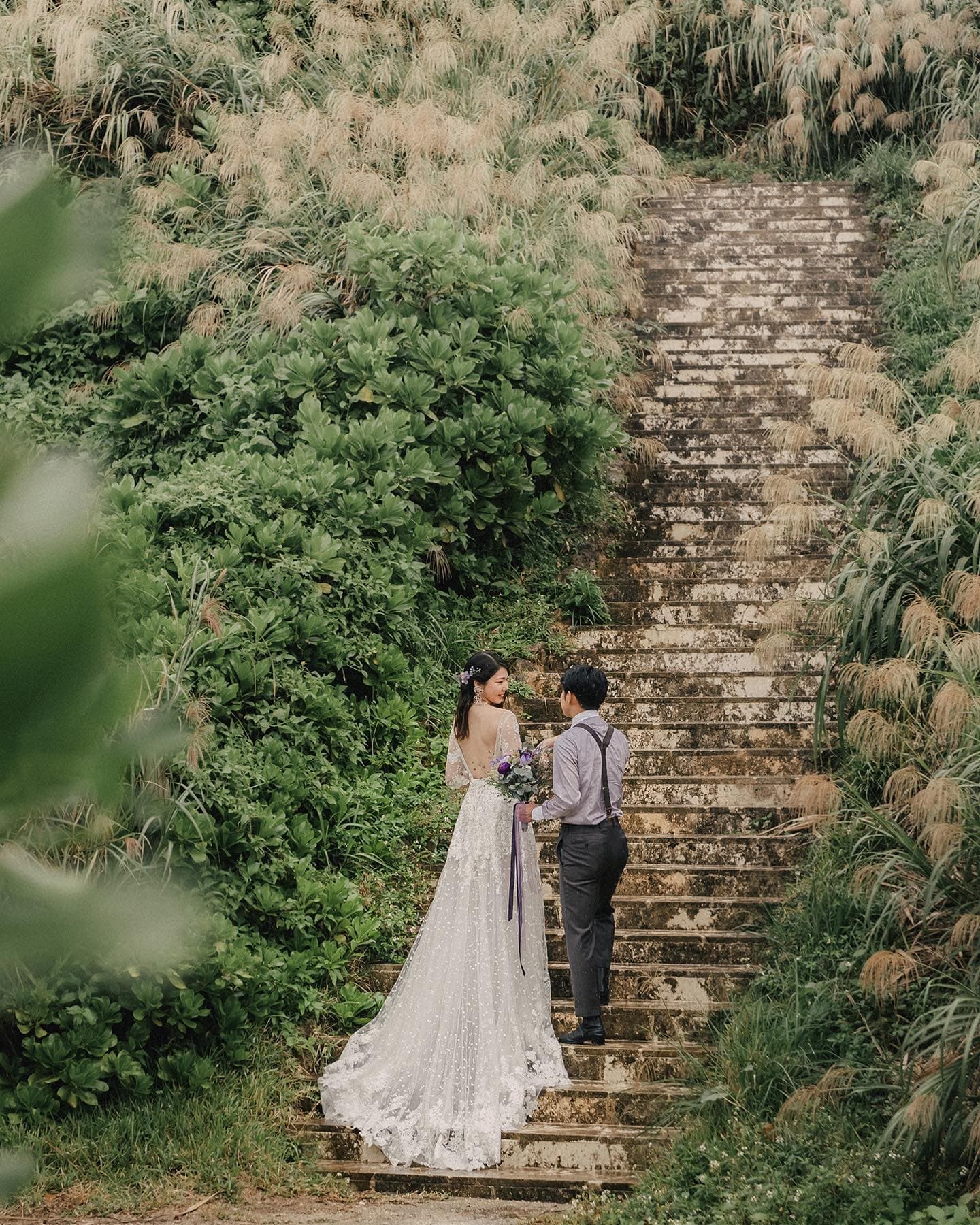Couple in wedding attire climbing an old stone stairway framed by pampas grass in Okinawa.