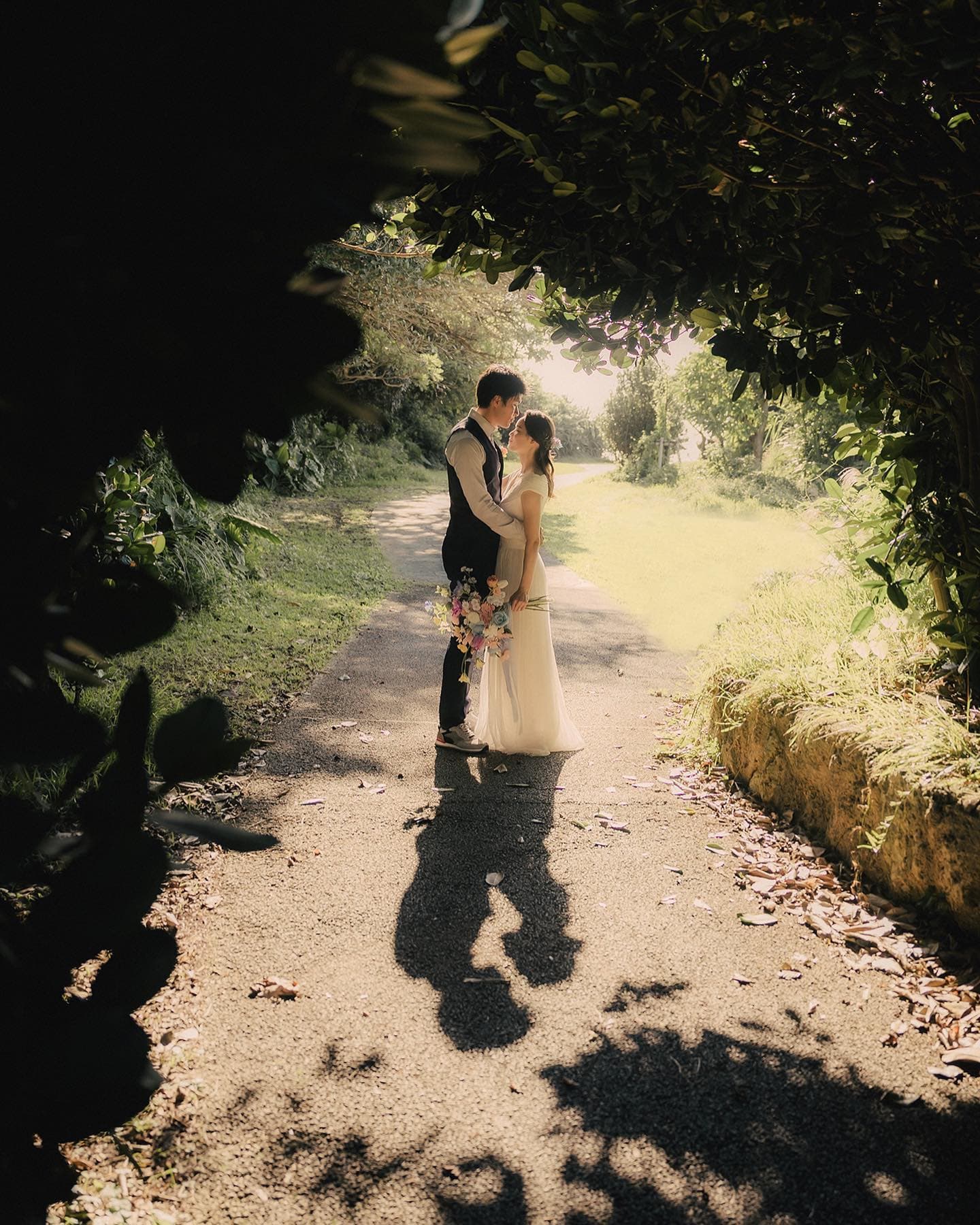 Couple kissing on a sun-dappled path framed by dark foliage in Okinawa