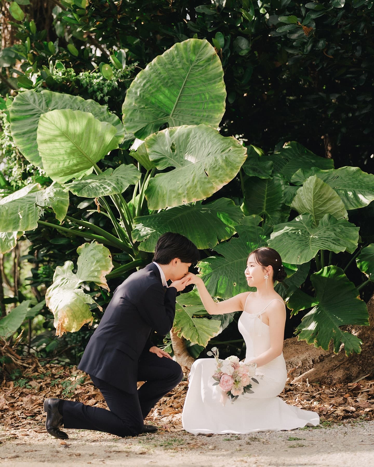 Groom kneeling to kiss the bride's hand before giant tropical leaves in Okinawa