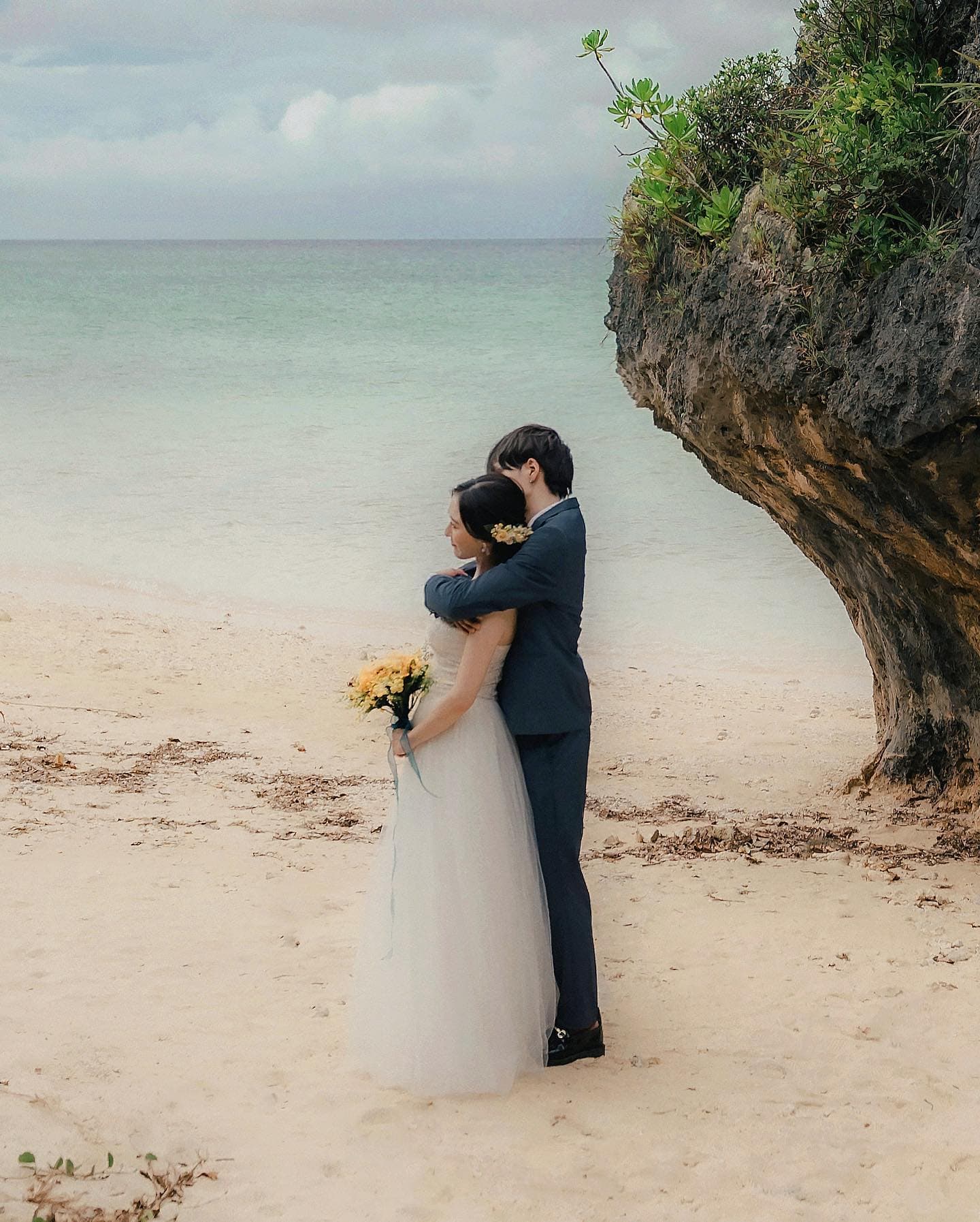 Couple embracing on a quiet Okinawa beach beside a rocky outcrop on a cloudy day