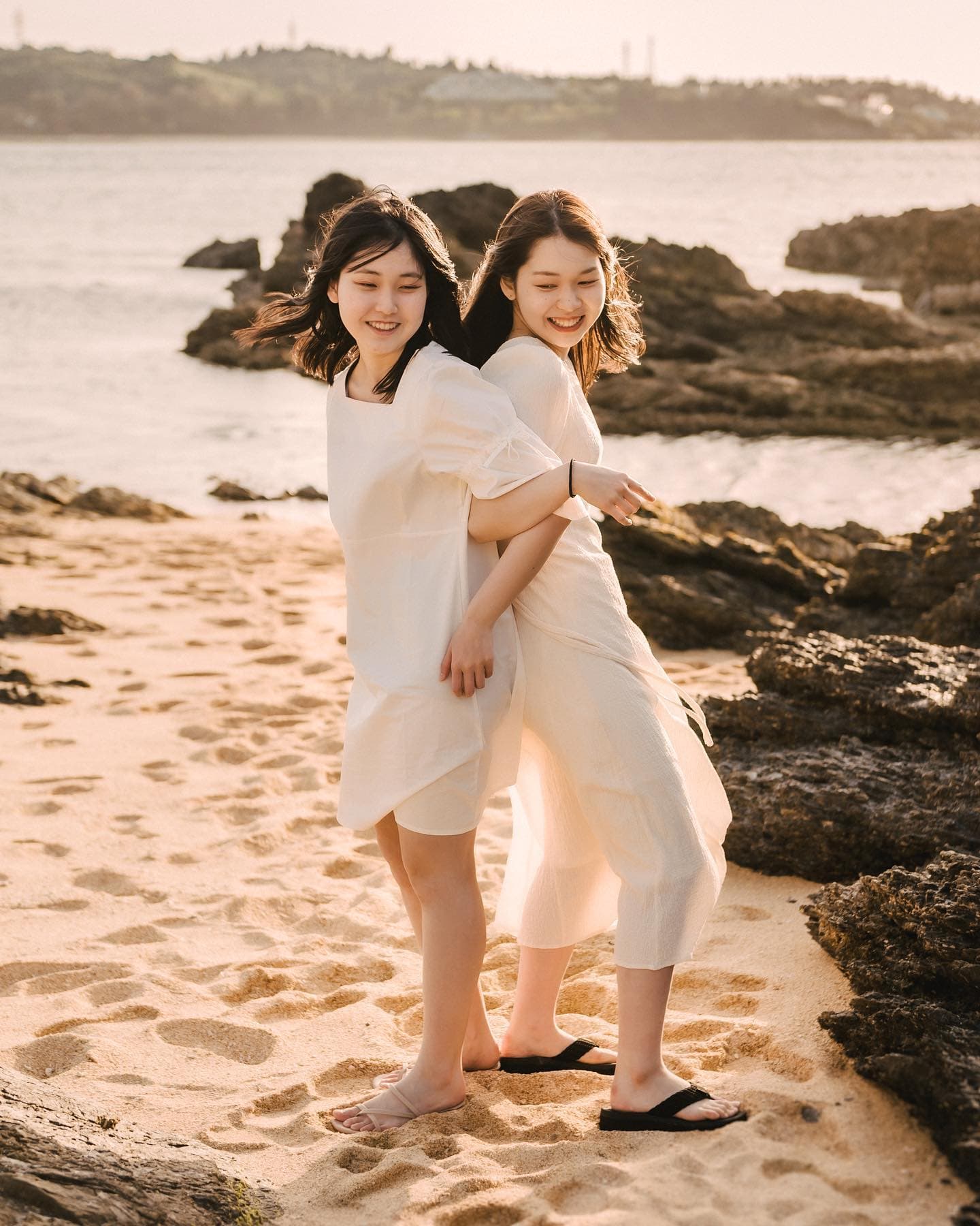 바위 해변에서 등을 맞대고 웃는 흰 원피스의 자매, 오키나와