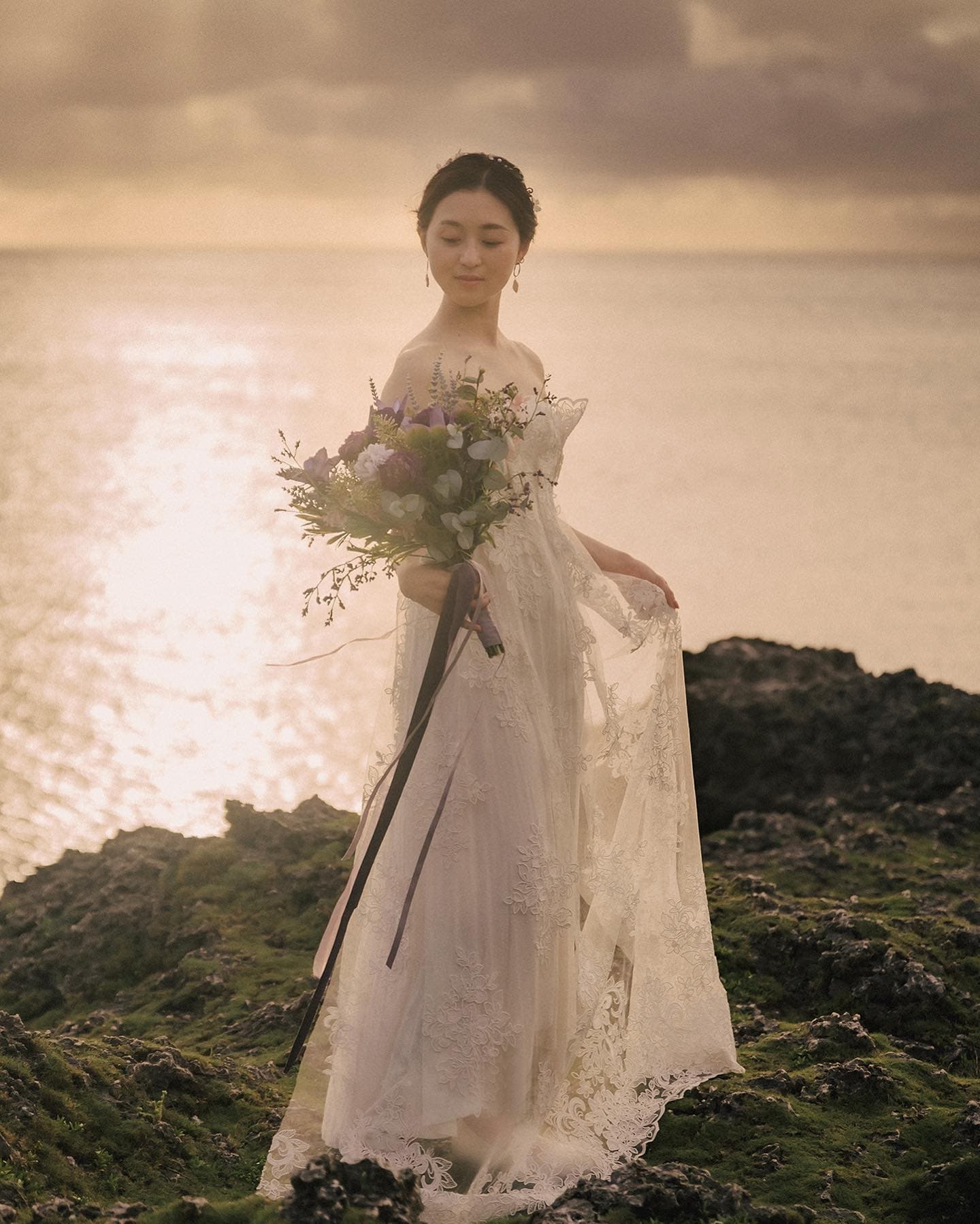 Bride in a lace gown holding her bouquet before a softly glowing Okinawa sea