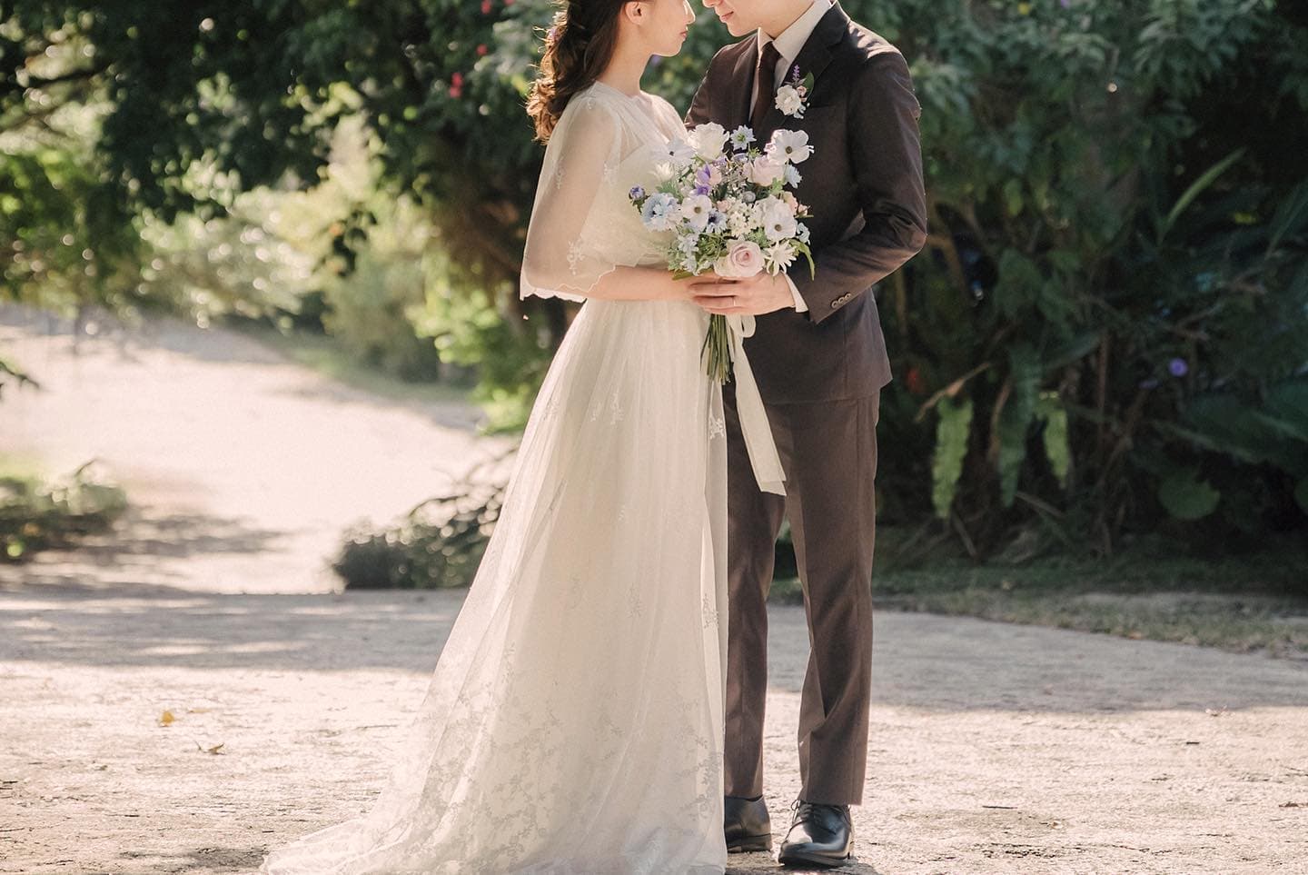 沖縄の並木道でブーケを挟んで向き合う白いドレスの花嫁と新郎