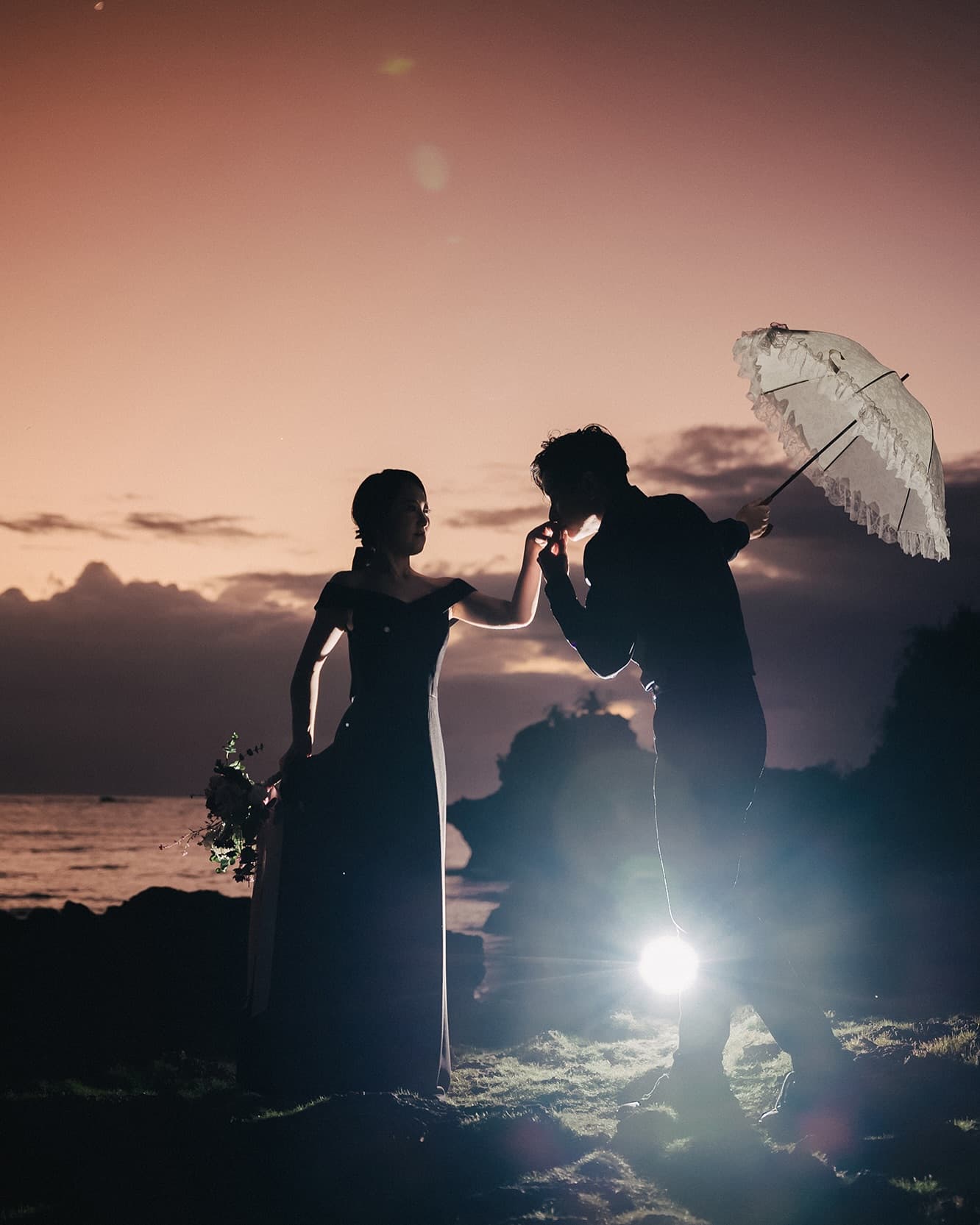 Groom kissing the bride's hand with a lace parasol at crimson Okinawa dusk