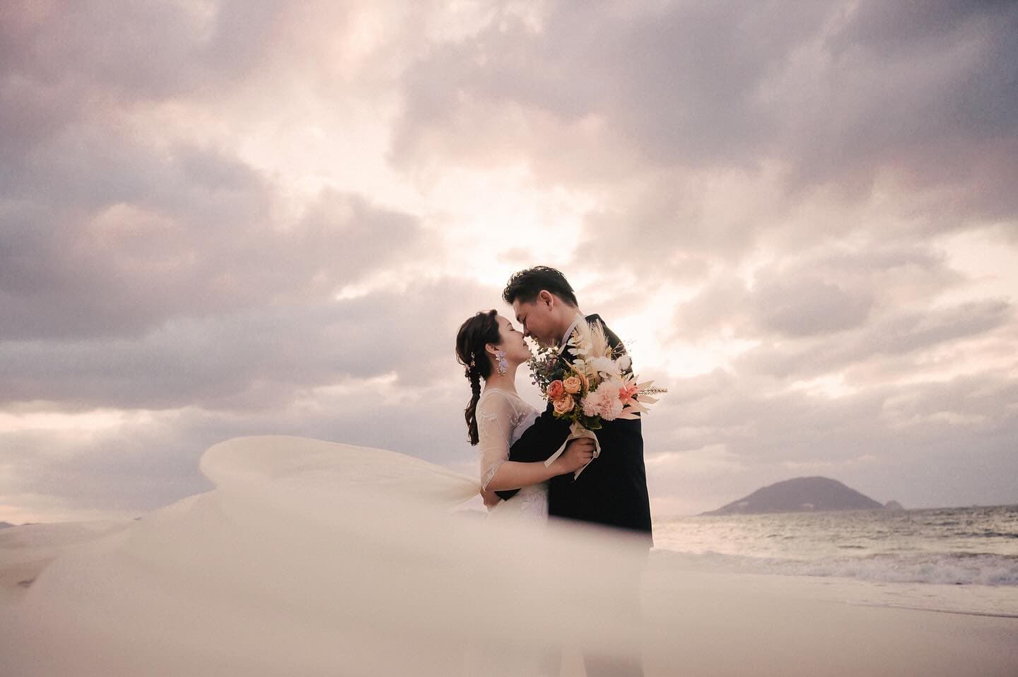 Bride and groom embracing on the shore, dress train flowing under a soft cloudy sky
