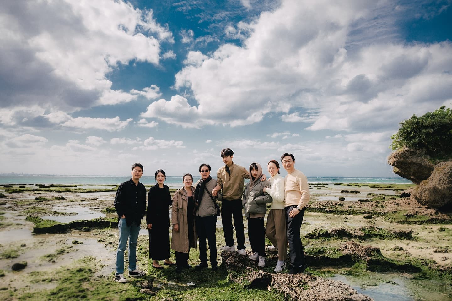 Large family posing on a rocky Okinawa shore under a cloudy blue sky