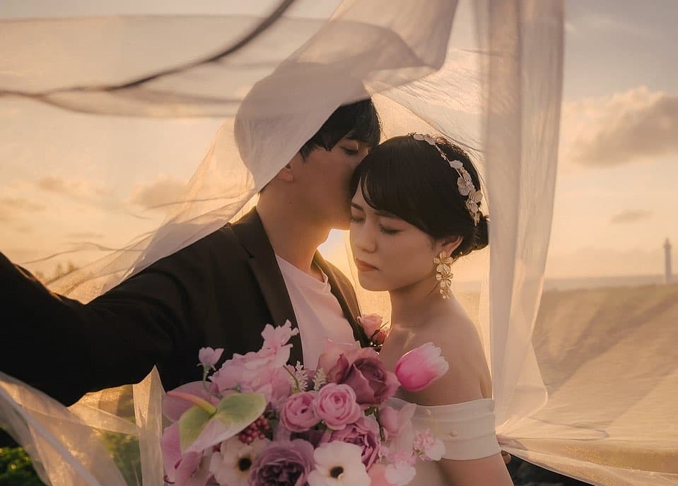 해 질 녘 오키나와 바닷가, 베일 아래 기댄 신랑 신부와 핑크 부케.