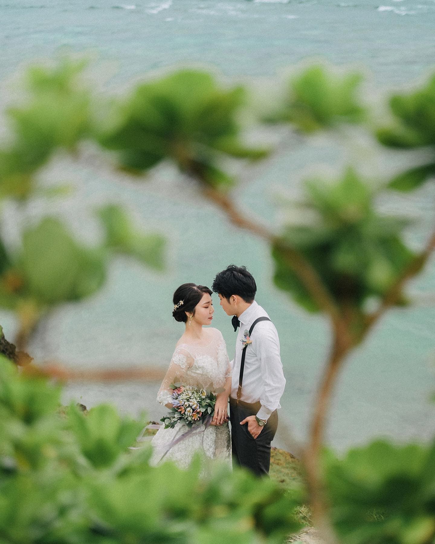 Through green foliage, a bride and groom face each other by the sea on an overcast day in Okinawa.