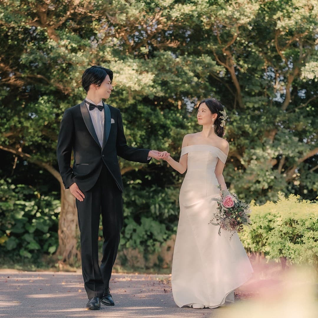 緑の並木道を手を取り歩く新郎新婦とピンクのブーケ。沖縄。